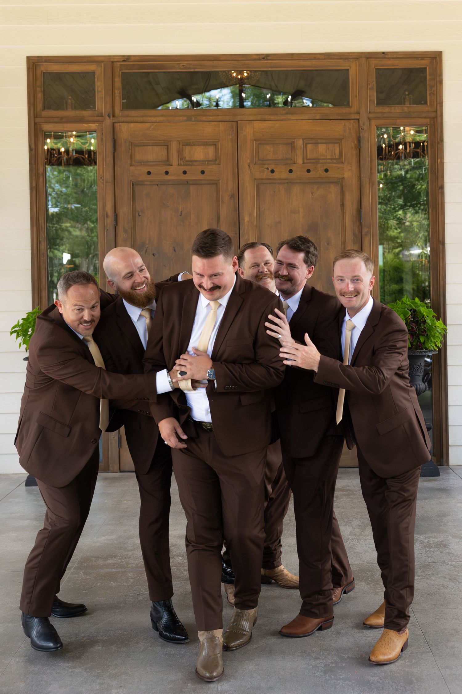 Groomsmen in brown suits strike playful domino-effect poses on a covered porch.
