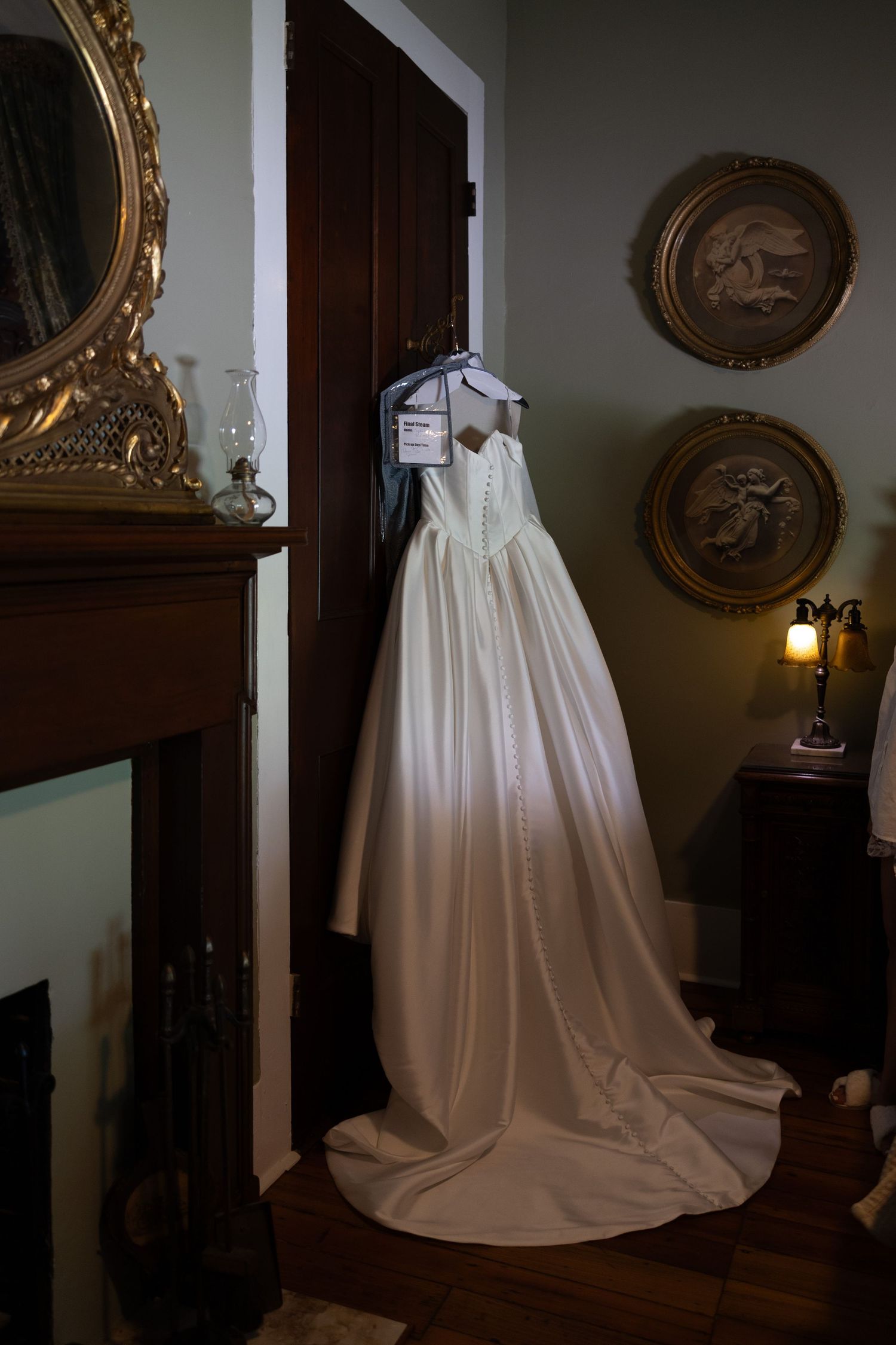 An elegant white wedding dress displayed in a dimly lit room with ornate gold-framed artwork on the wall.