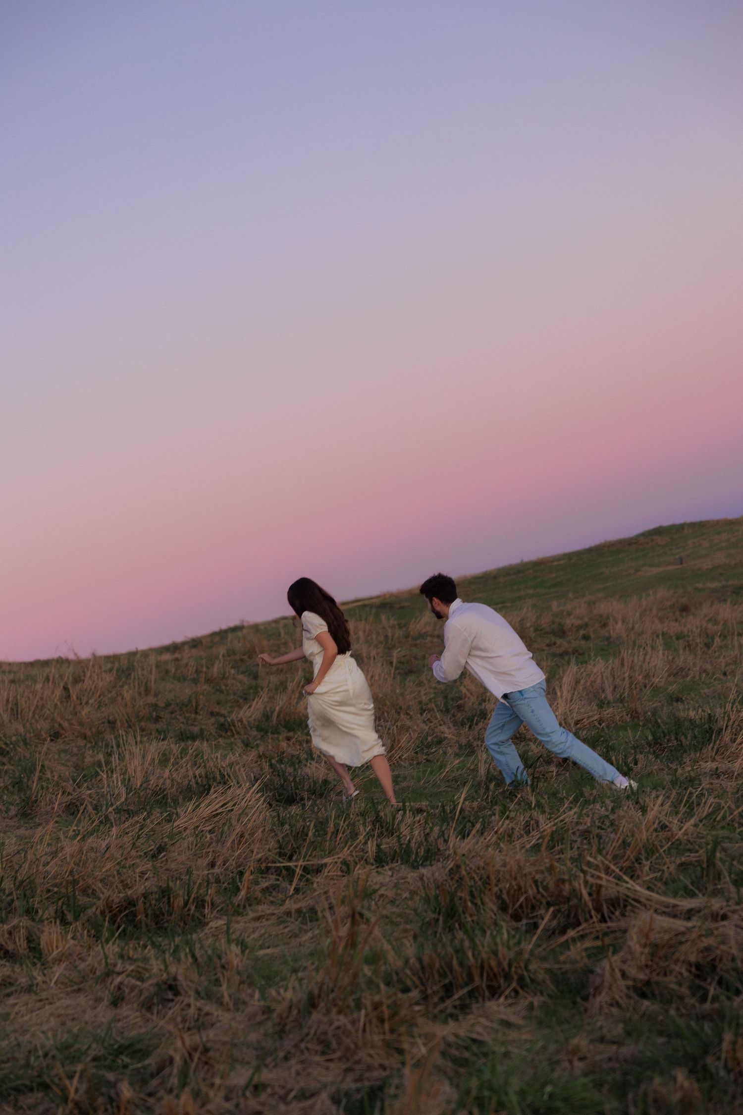 Two people climb up a grassy hillside at sunset with a pink and purple sky in the background.