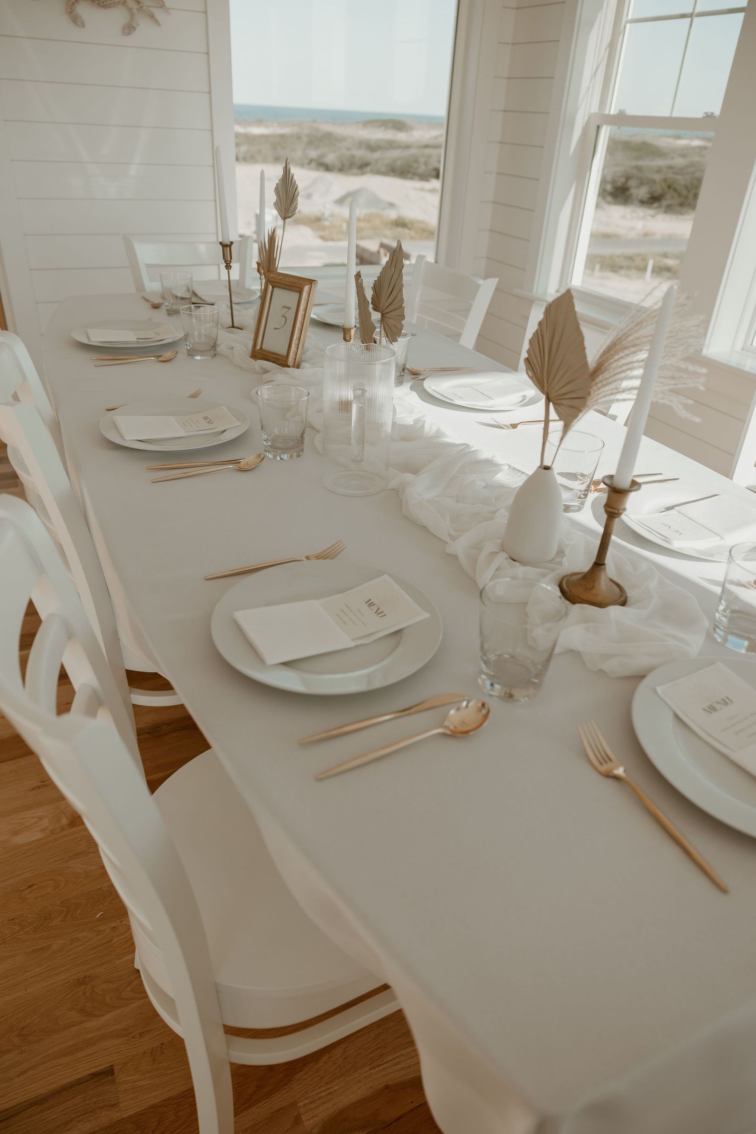 Elegant white tablescape with place settings and coastal view through windows.