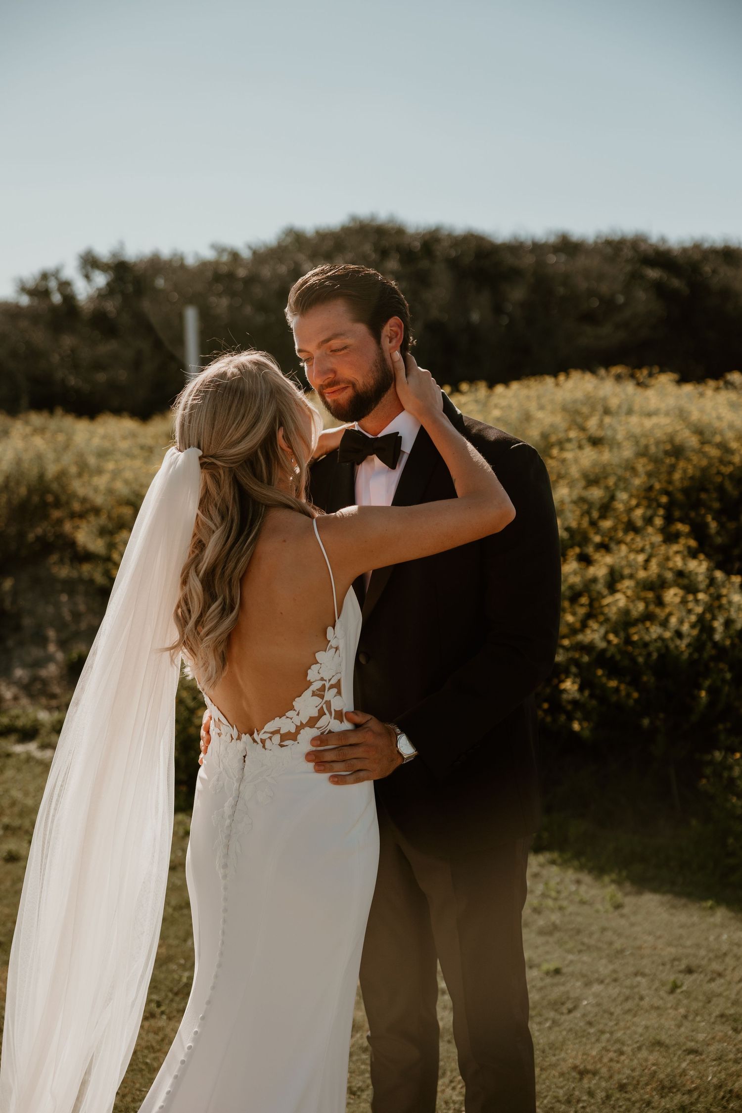 A bride and groom share an intimate moment in a sun-lit outdoor setting with natural foliage in the background.