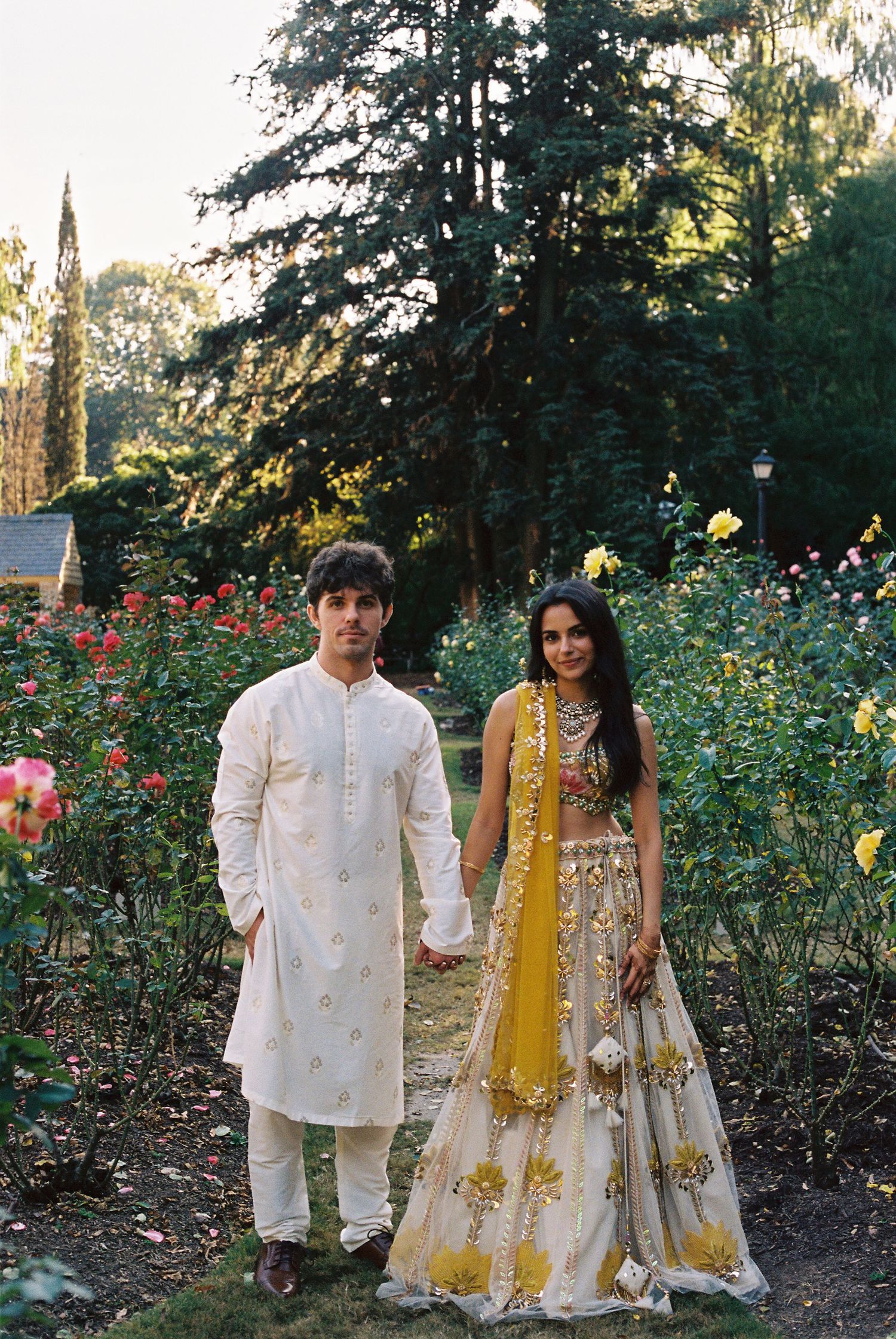A couple in traditional Indian wedding attire stands together in a garden of roses.