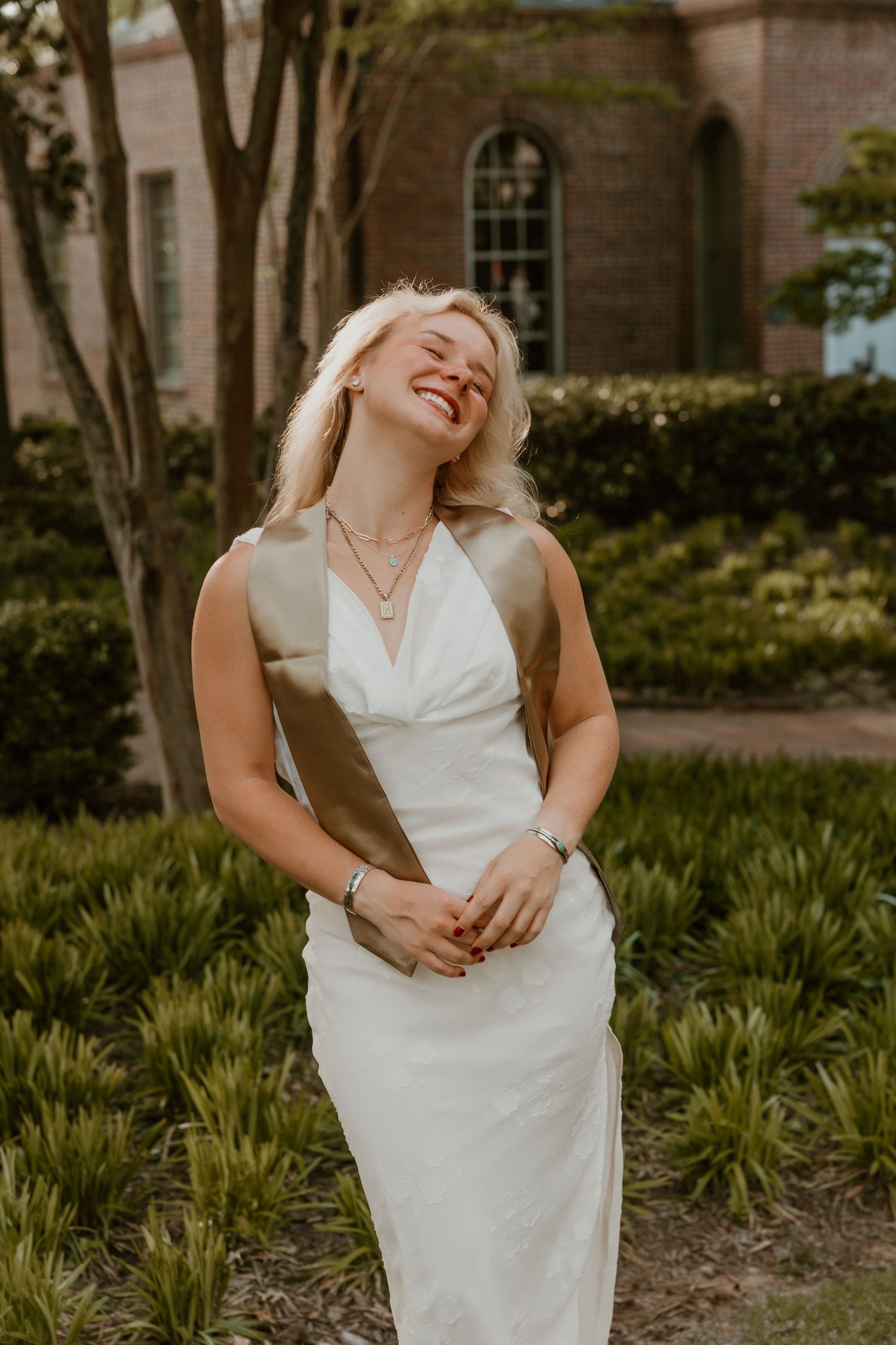 Smiling blonde woman in white halter gown with gold sash laughing outdoors near brick building and lush greenery.