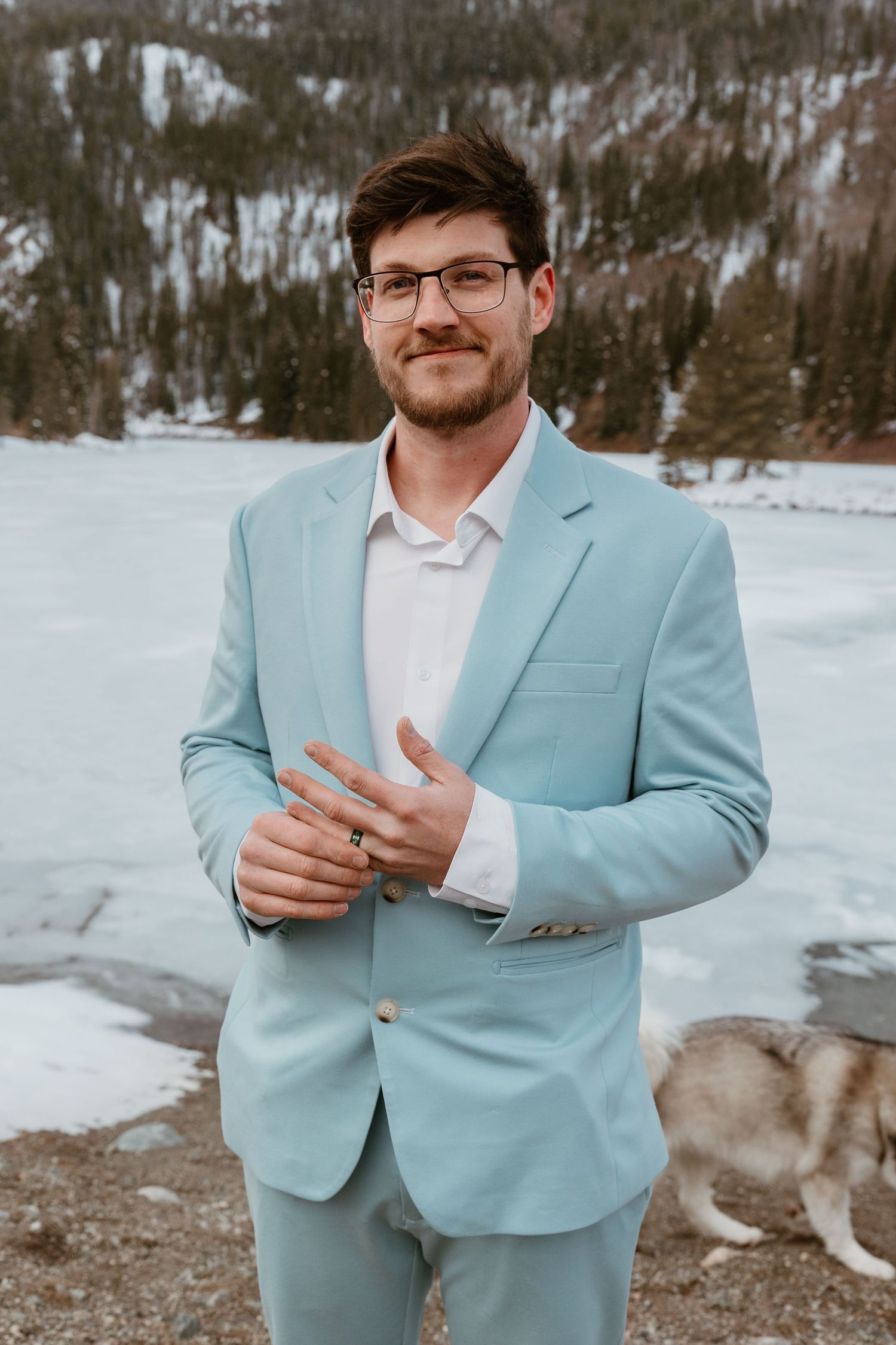 A person wearing a light blue suit and glasses stands outdoors in a snowy mountain setting.