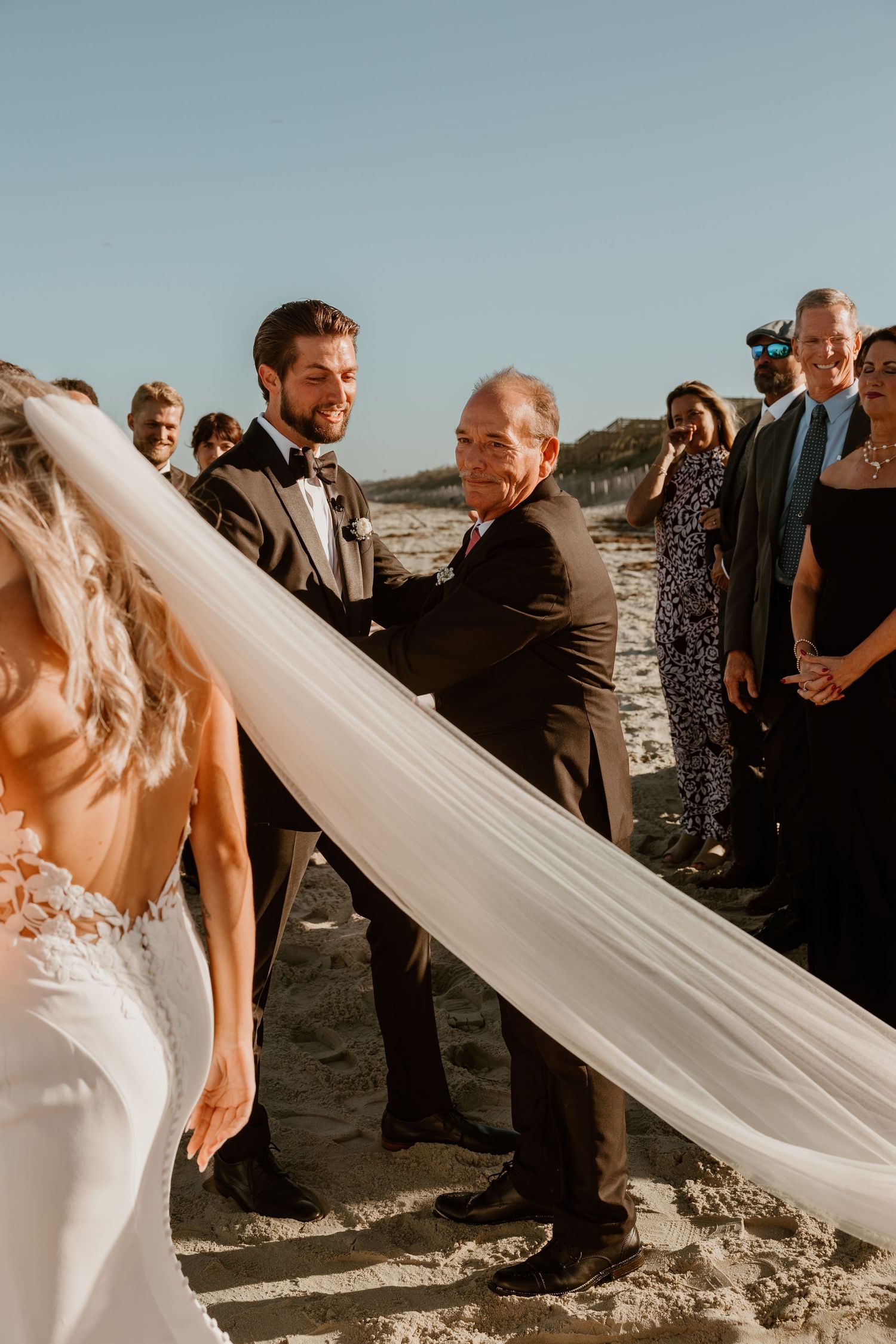 Beach wedding ceremony with flowing white dress and guests gathered on sandy shore.