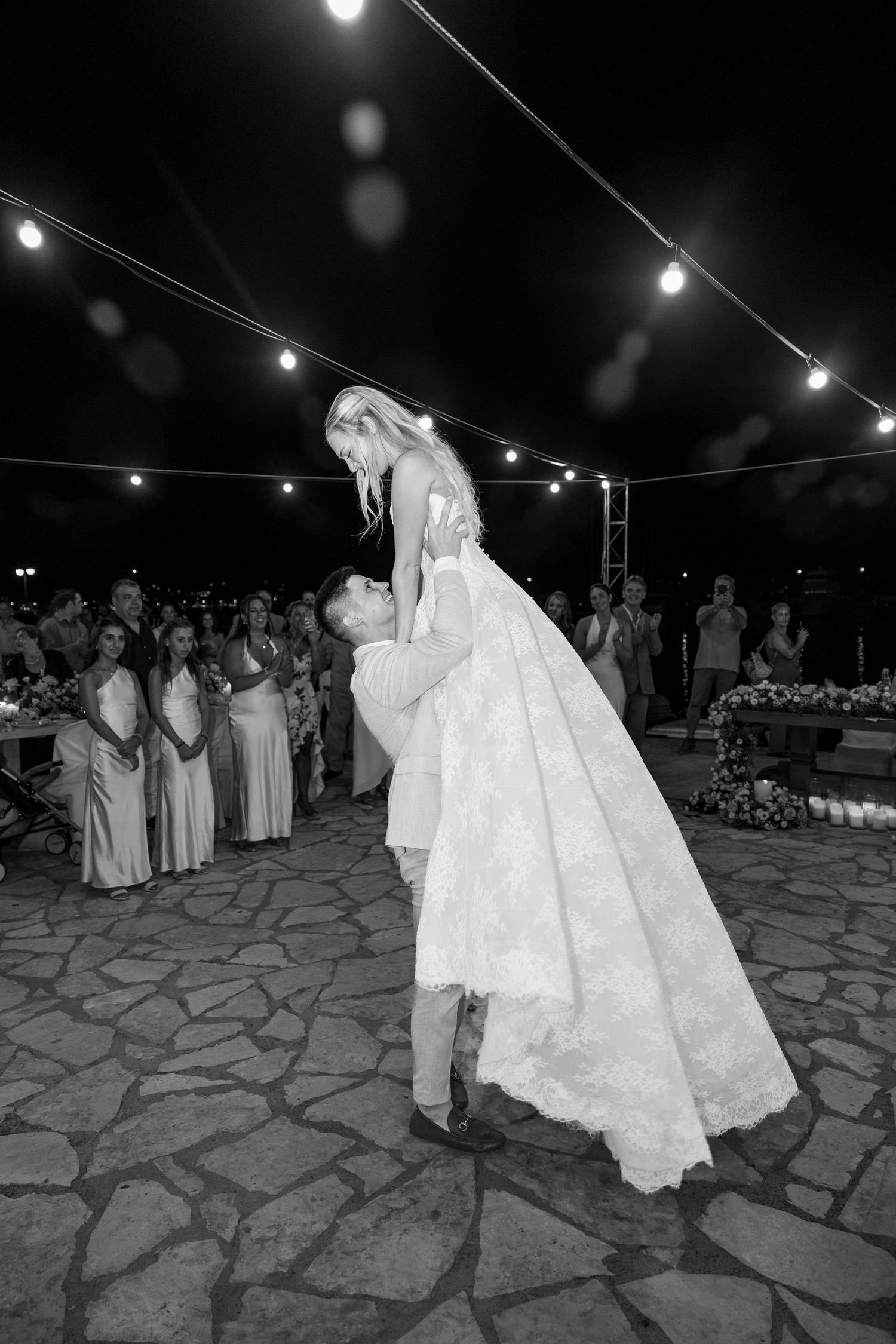 A couple shares a romantic dance on a stone patio under string lights at an outdoor evening wedding reception.
