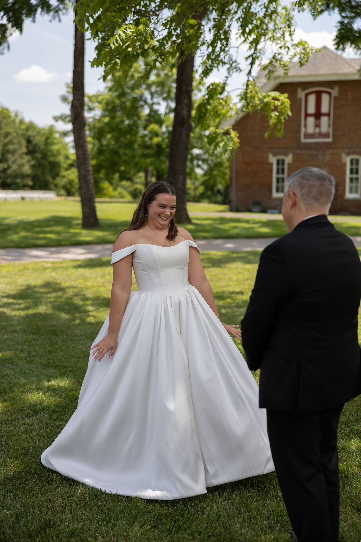 A couple in wedding attire walking together on a sunny lawn with brick buildings in the background.