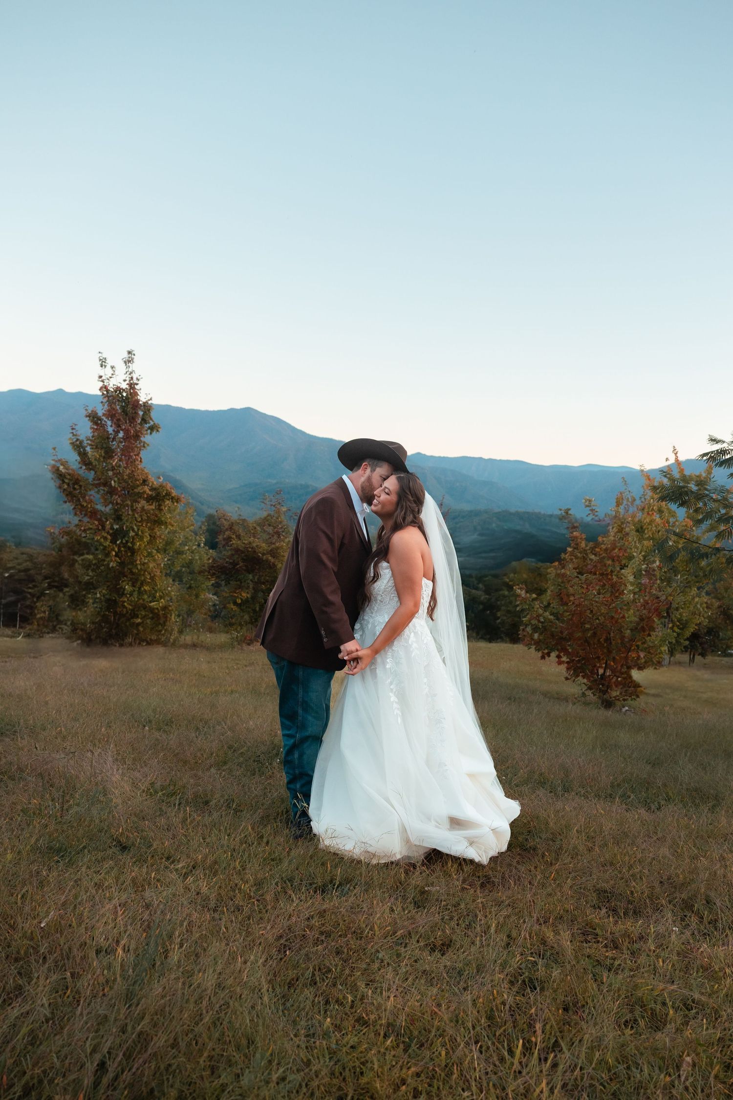 A couple in wedding attire embracing on a hilltop with mountains and autumn trees in the background.