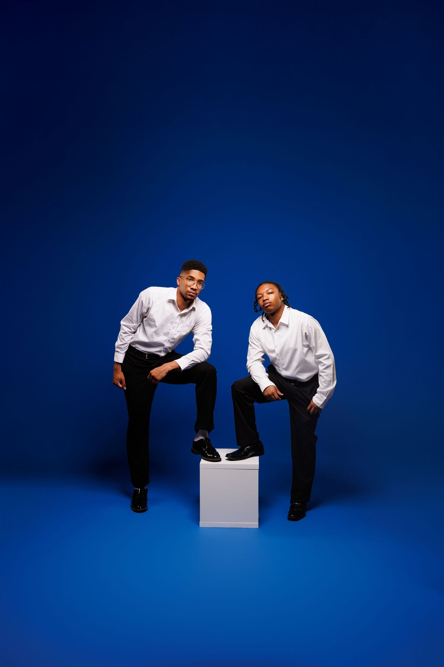 Studio portrait of two people in white button-downs posing with a white cube against blue background.