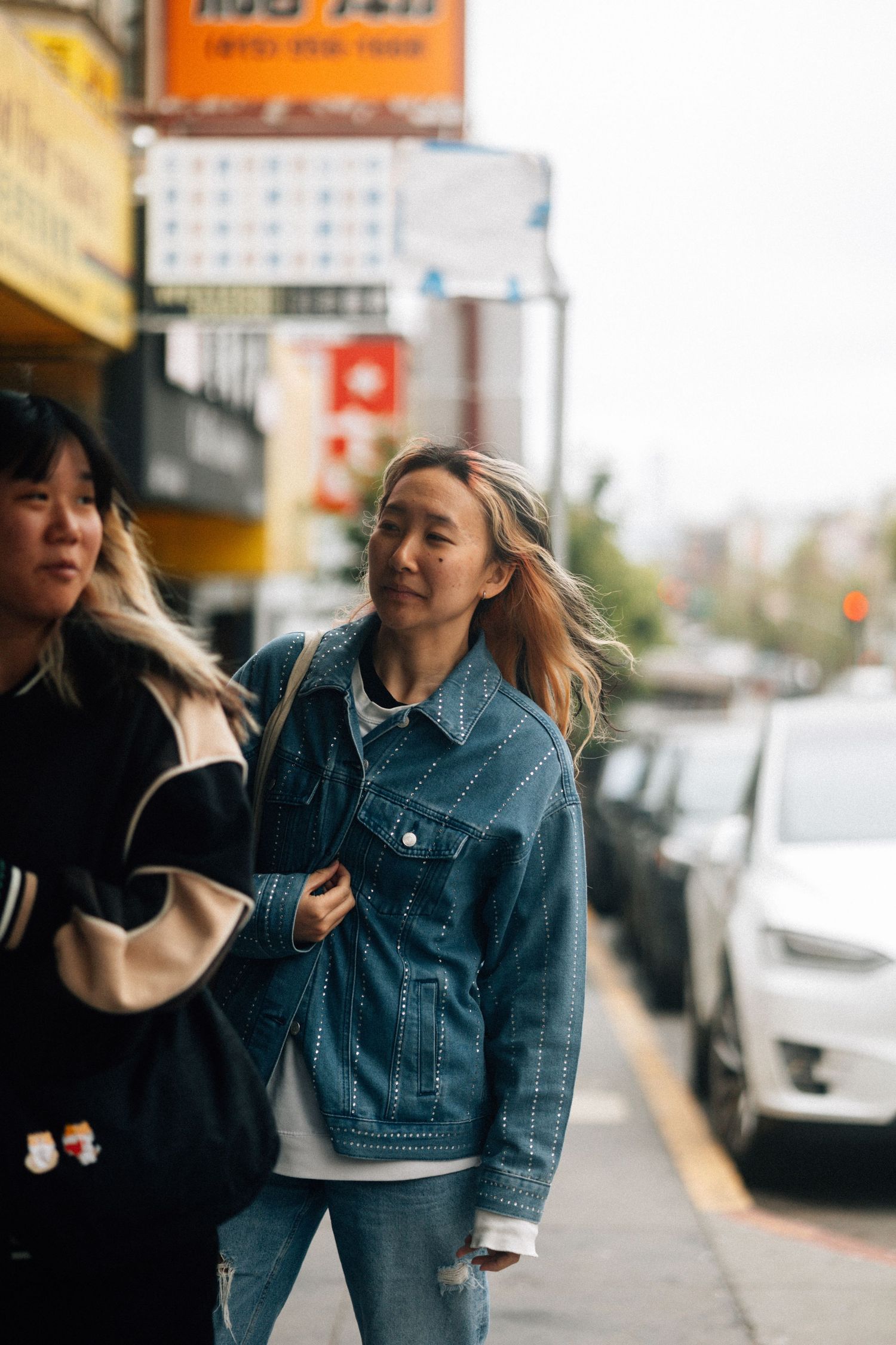 Two people walking down an urban sidewalk on a cloudy day, one wearing a denim jacket and the other in black outerwear.