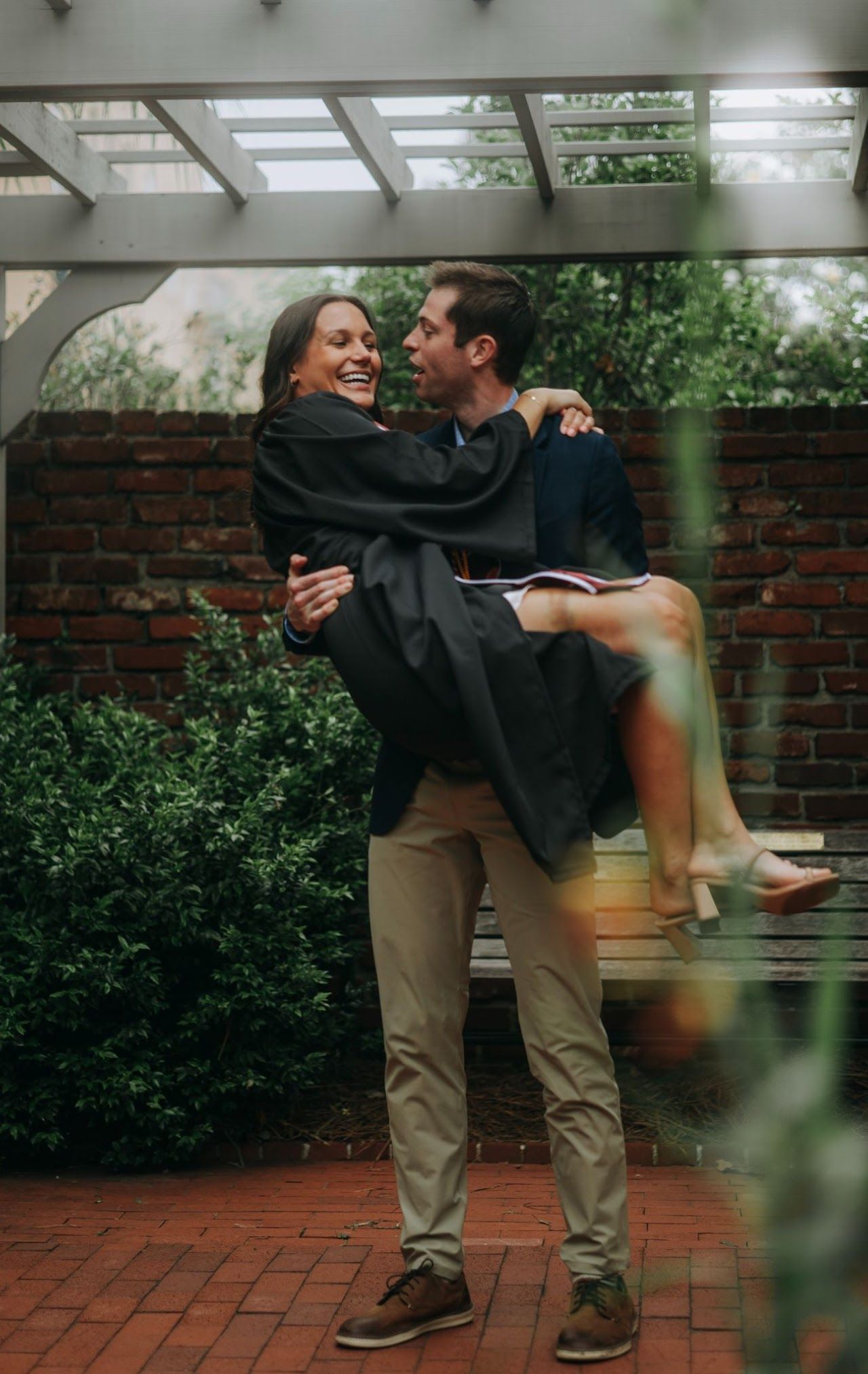 Couple in graduation attire sharing a joyful moment under a garden pergola during commencement celebrations.