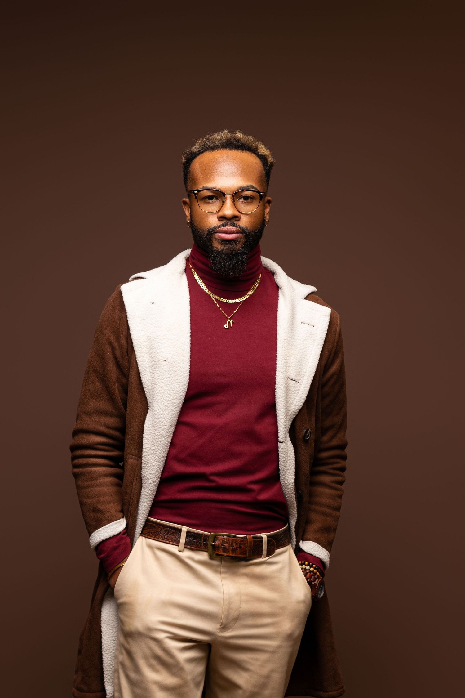 Stylish portrait of man in burgundy sweater and brown coat against dark background.