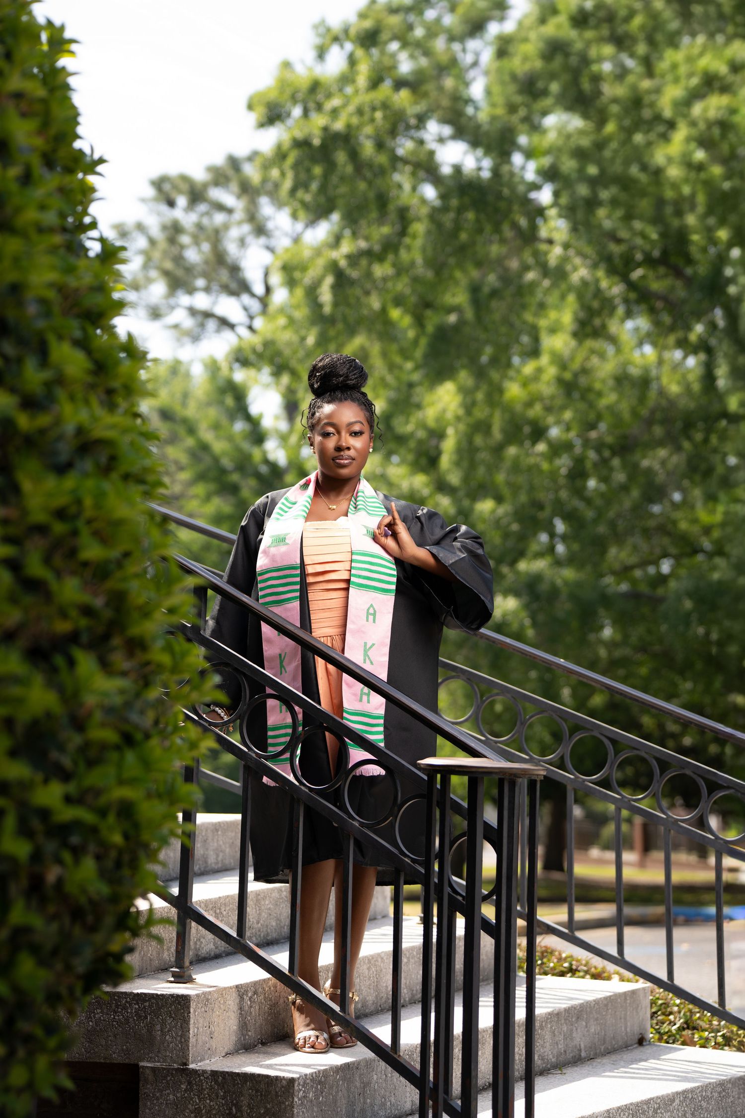 A young Black woman in graduation cap and gown with colorful stole poses proudly on stone steps outdoors.