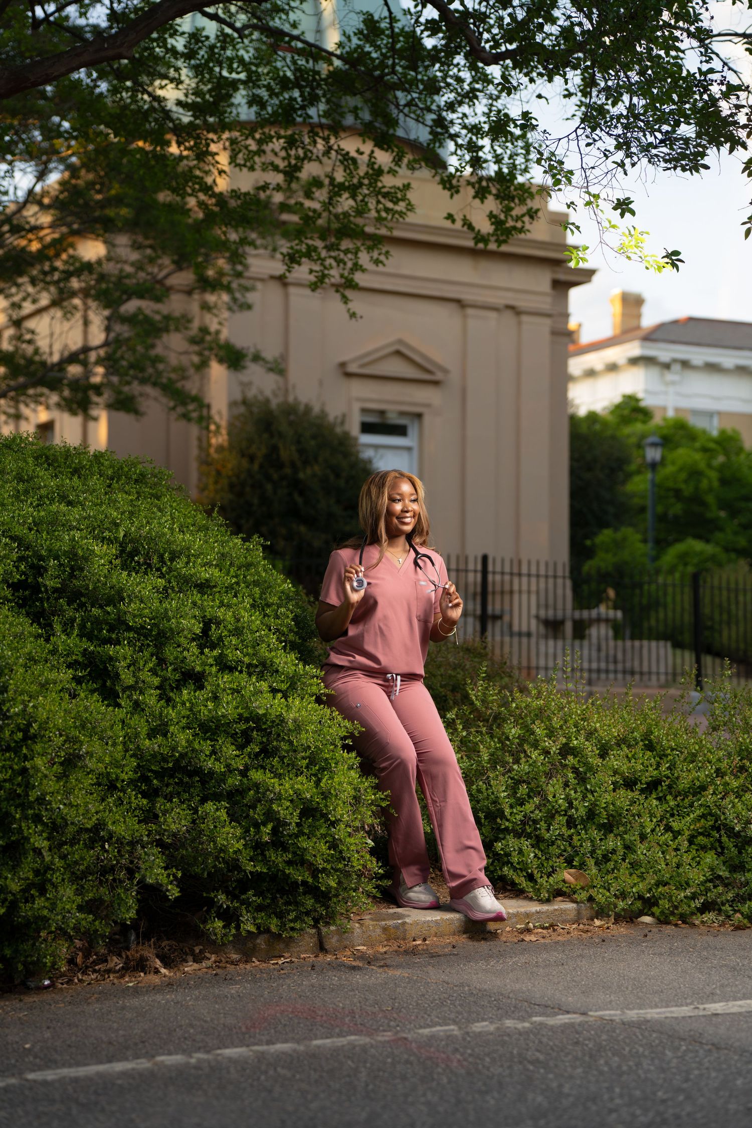 Young woman in pink scrubs with stethoscope poses outdoors near green bushes beside a tan building.