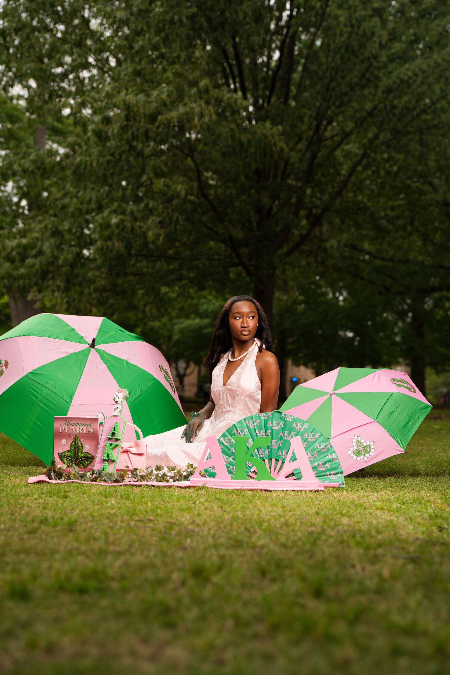 Young Black woman in white dress poses outdoors with pink and green umbrellas and fan on grass near trees.