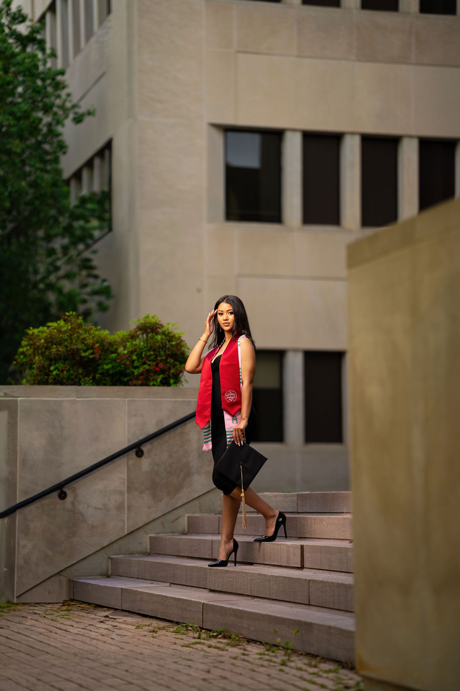 Graduate in red stole carries cap down outdoor stone steps near university building.
