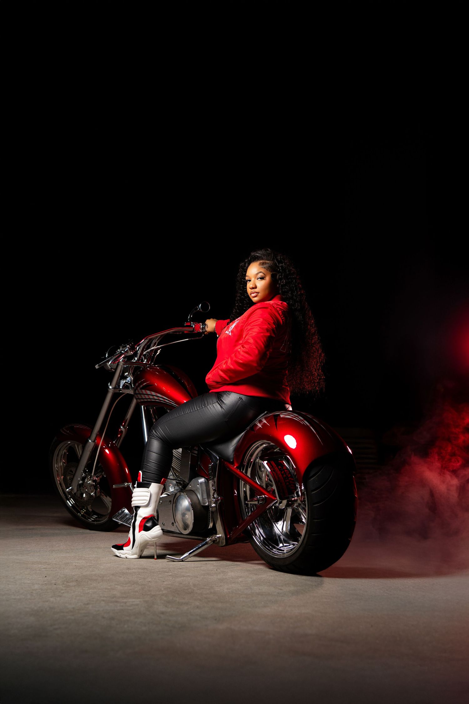 A woman in a red jacket and leather pants poses on a red custom chopper motorcycle against a dark smoky background.
