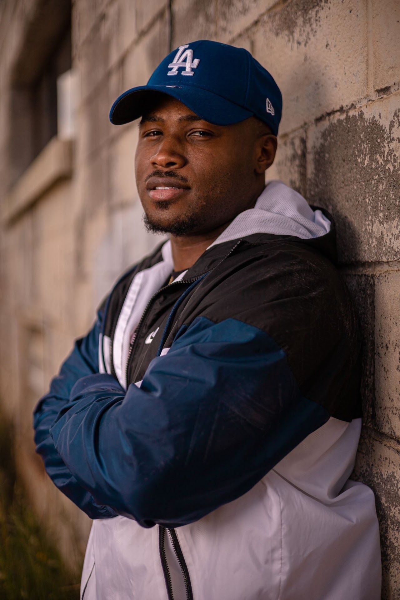 Person wearing LA Dodgers cap and white hoodie leans against wall in outdoor portrait setting.