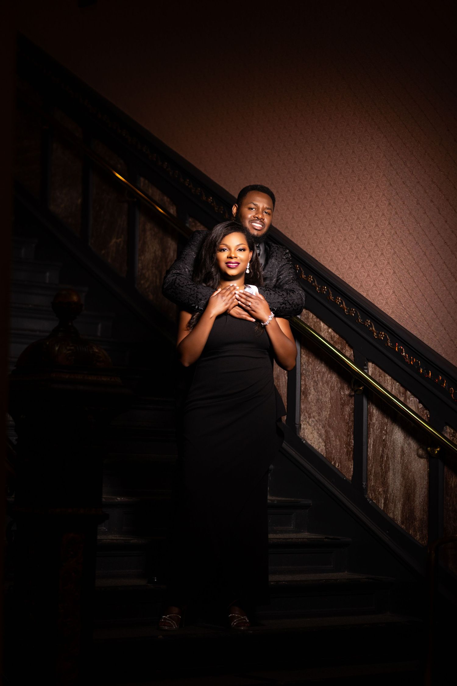 Romantic couple pose on dimly lit staircase in formal evening wear.