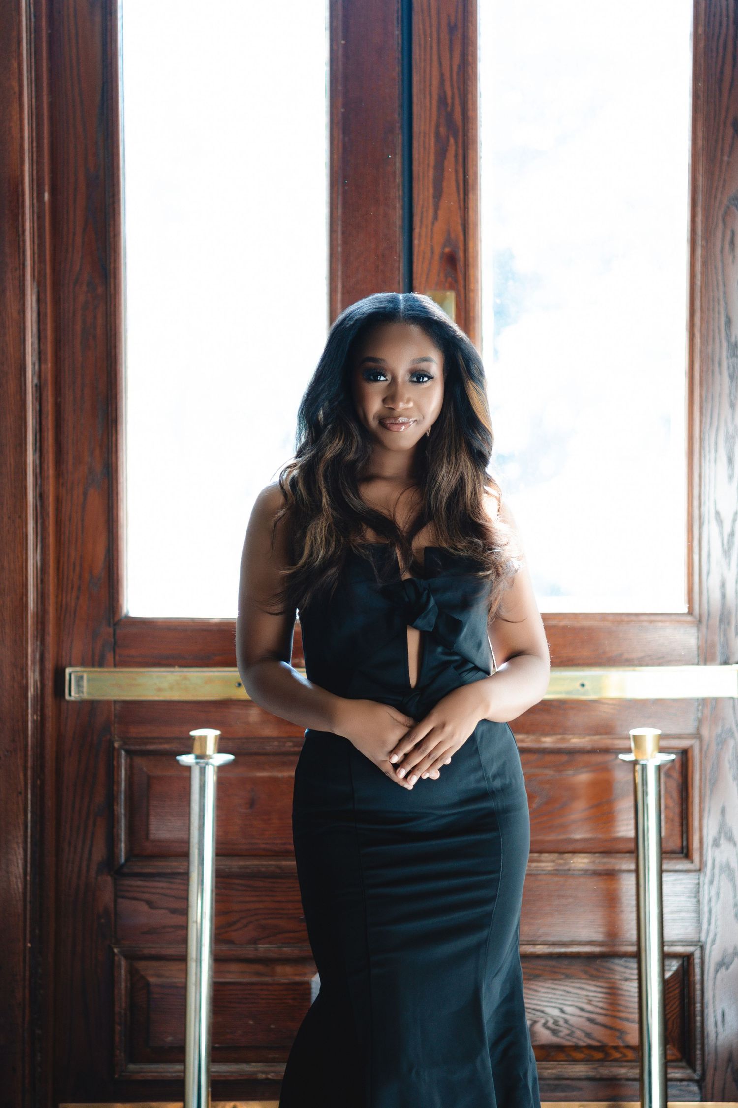 Elegant poses in black dress near wooden windows with natural lighting.