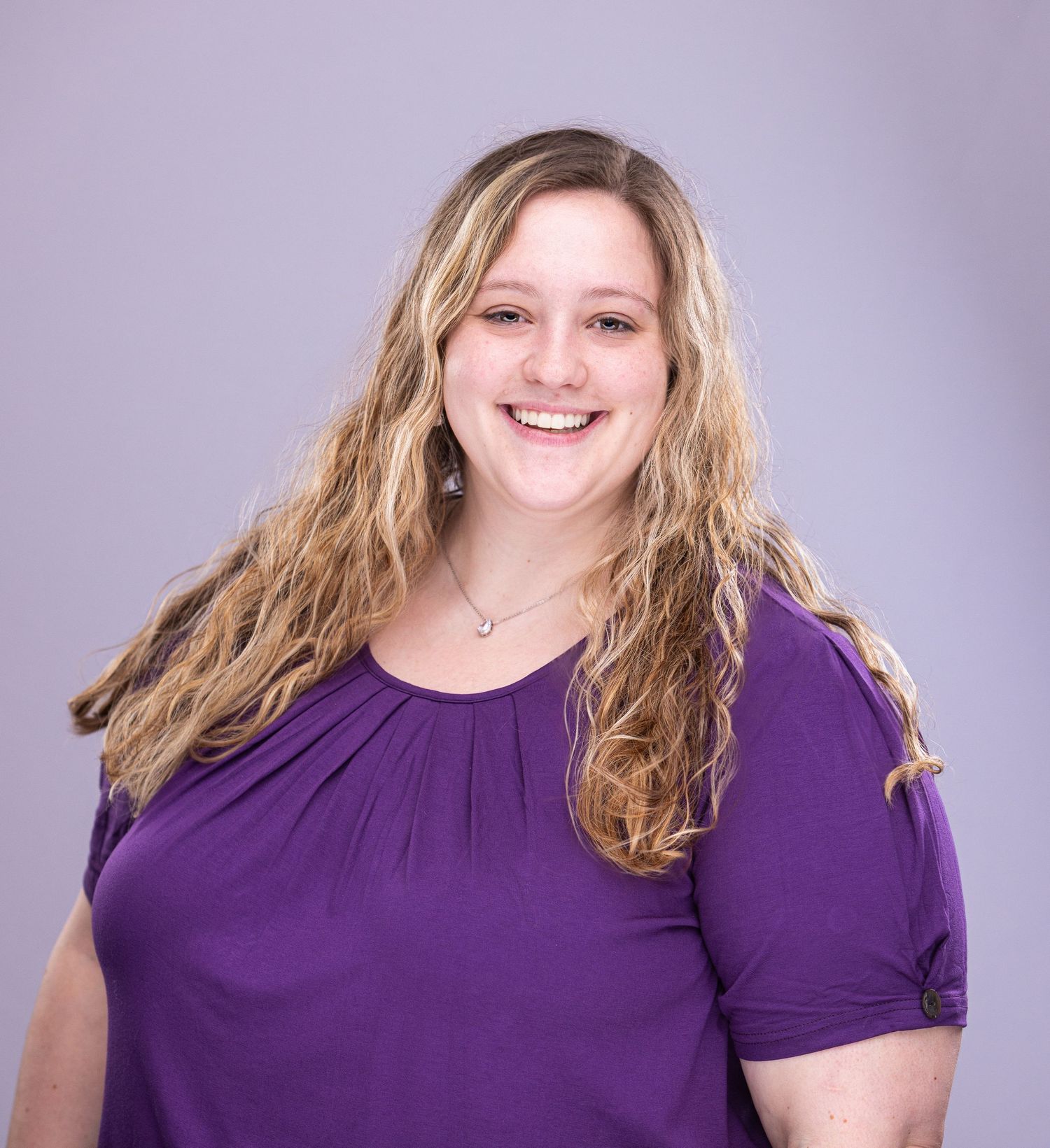Cheerful portrait of person in purple top with wavy blonde hair against light background.