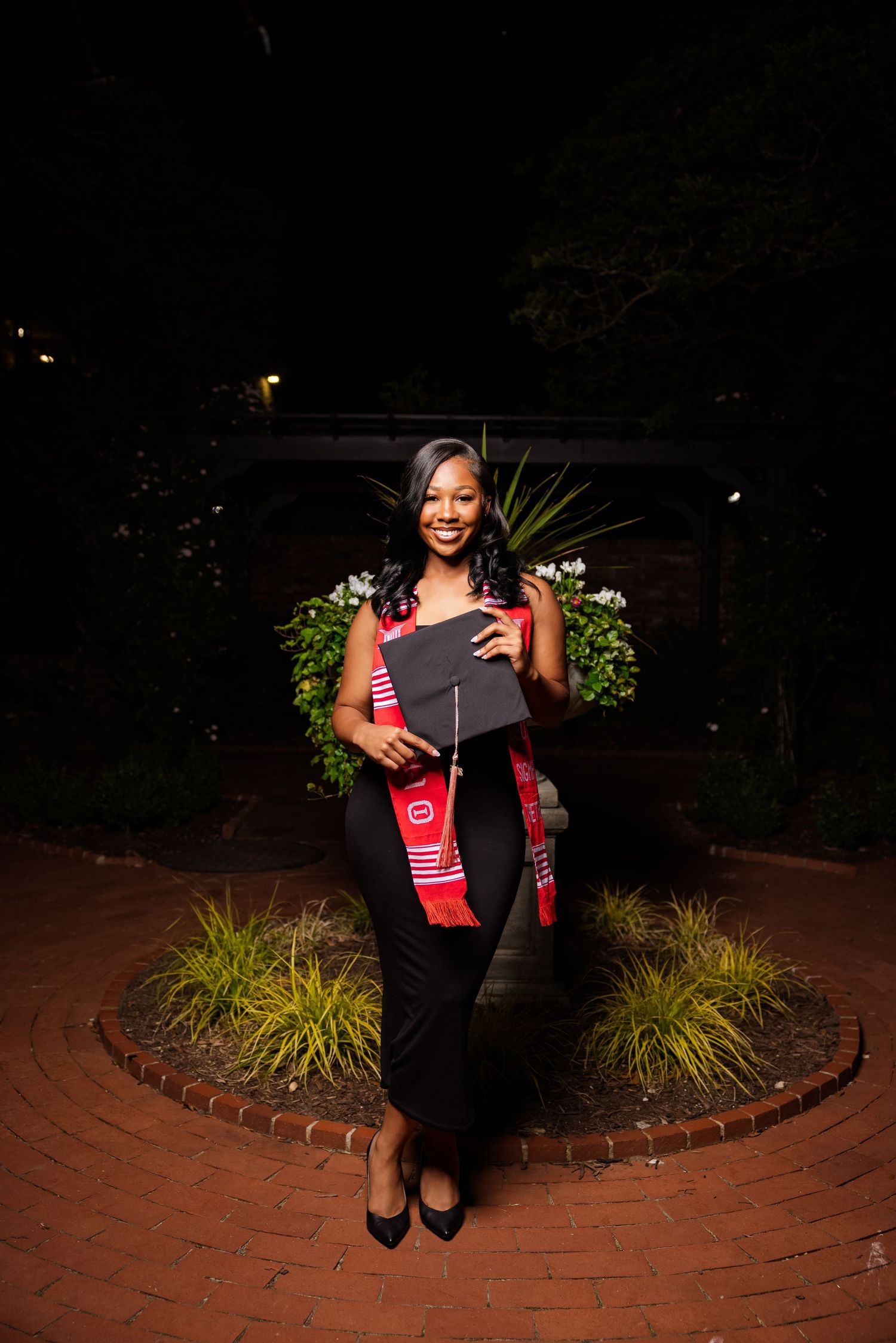 Graduate holding diploma at night in front of landscaped brick pathway.