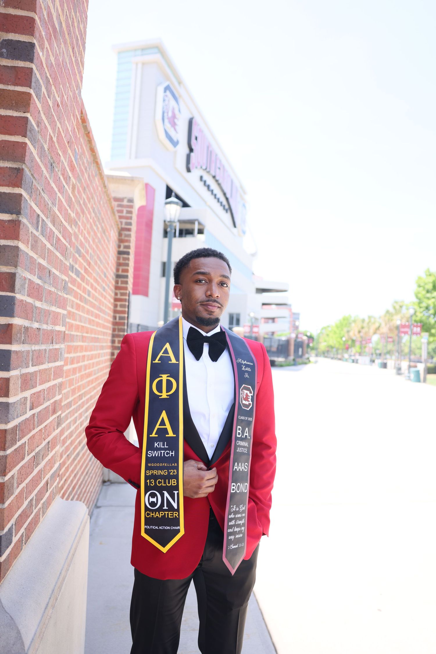 Graduate in red tuxedo jacket and fraternity stoles poses against brick wall.