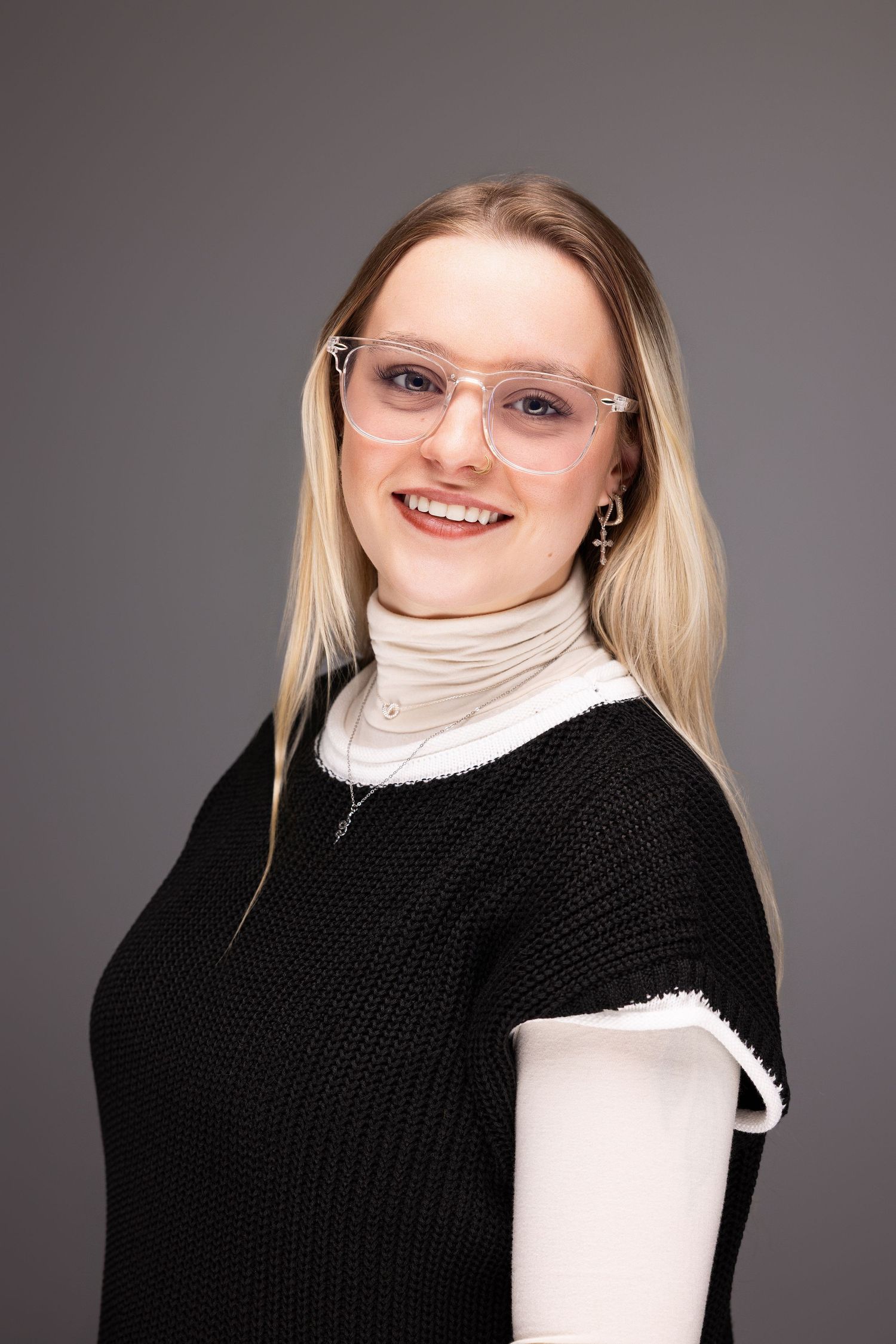 Professional studio portrait showing person in black sweater and white turtleneck with glasses against gray backdrop.