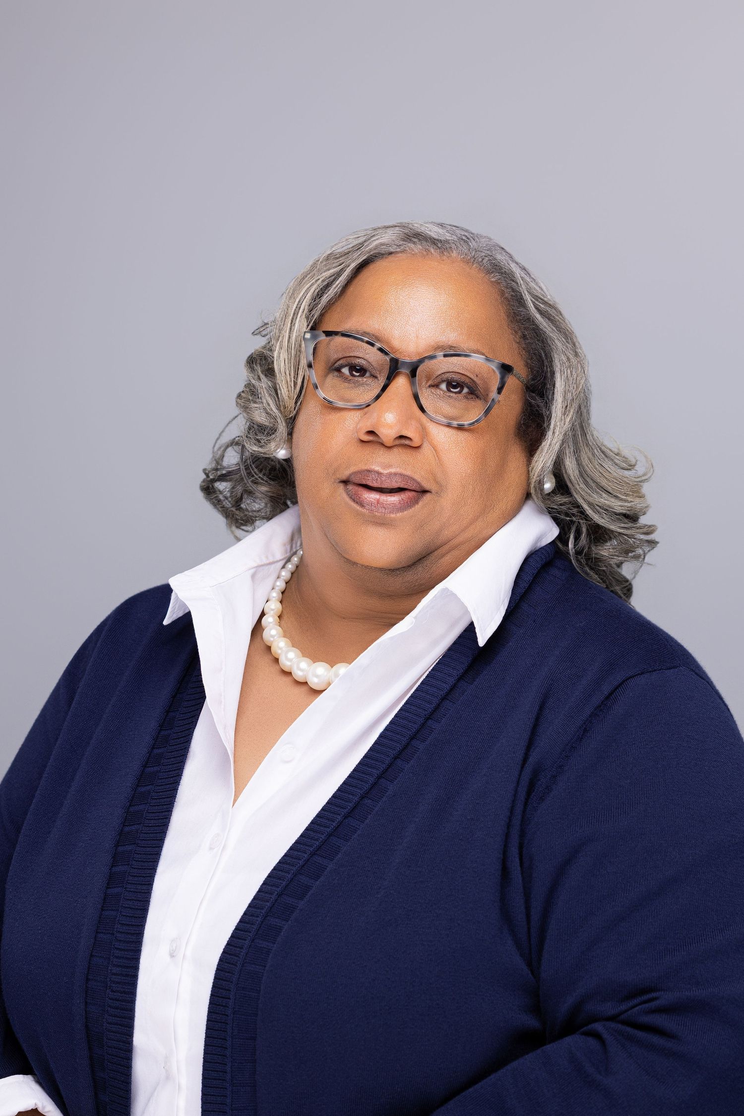 Professional headshot of person wearing glasses, a white collared shirt and navy cardigan with pearl necklace against gray backdrop.