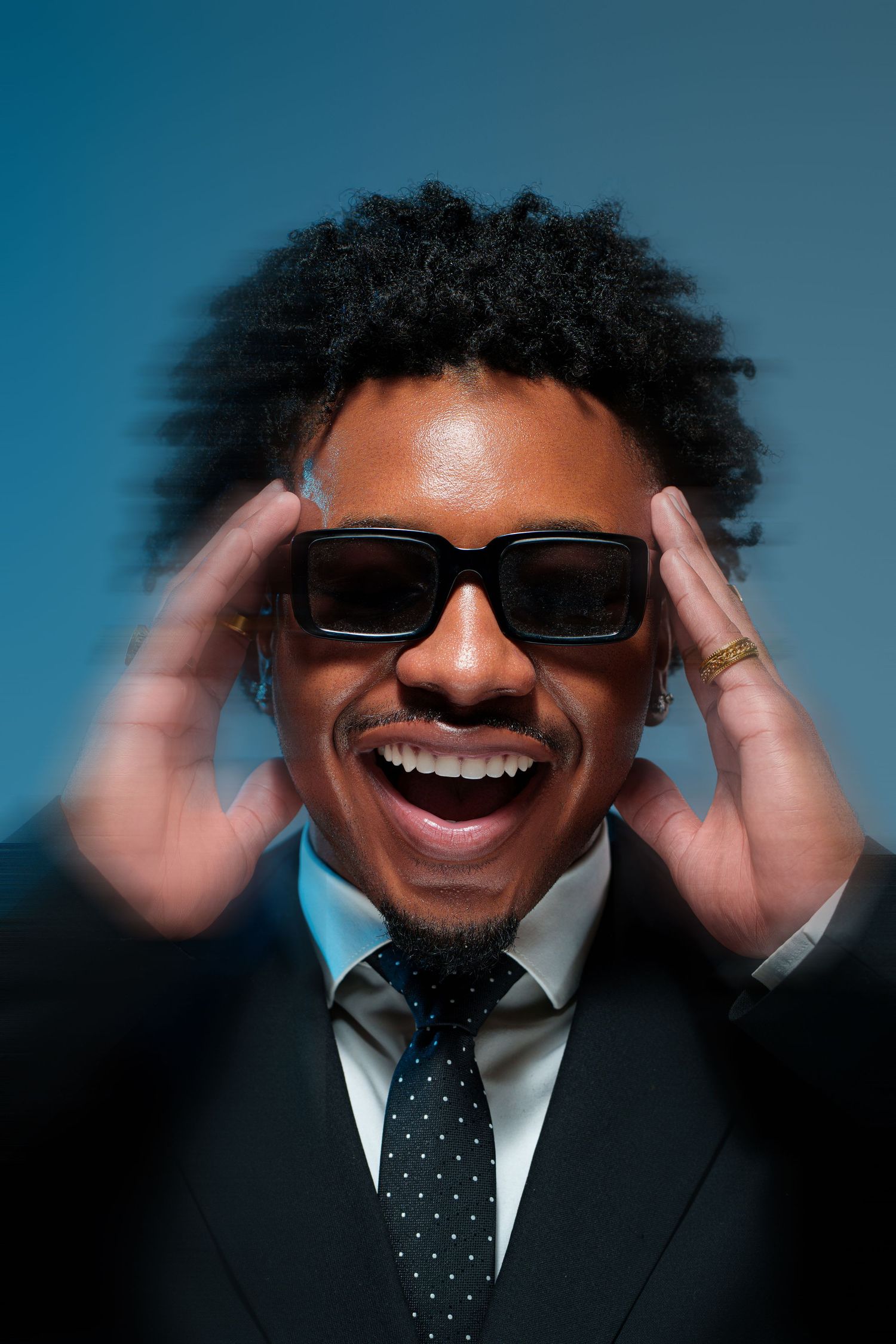 A smiling Black man in a suit and tie adjusts his dark sunglasses against a blue background.