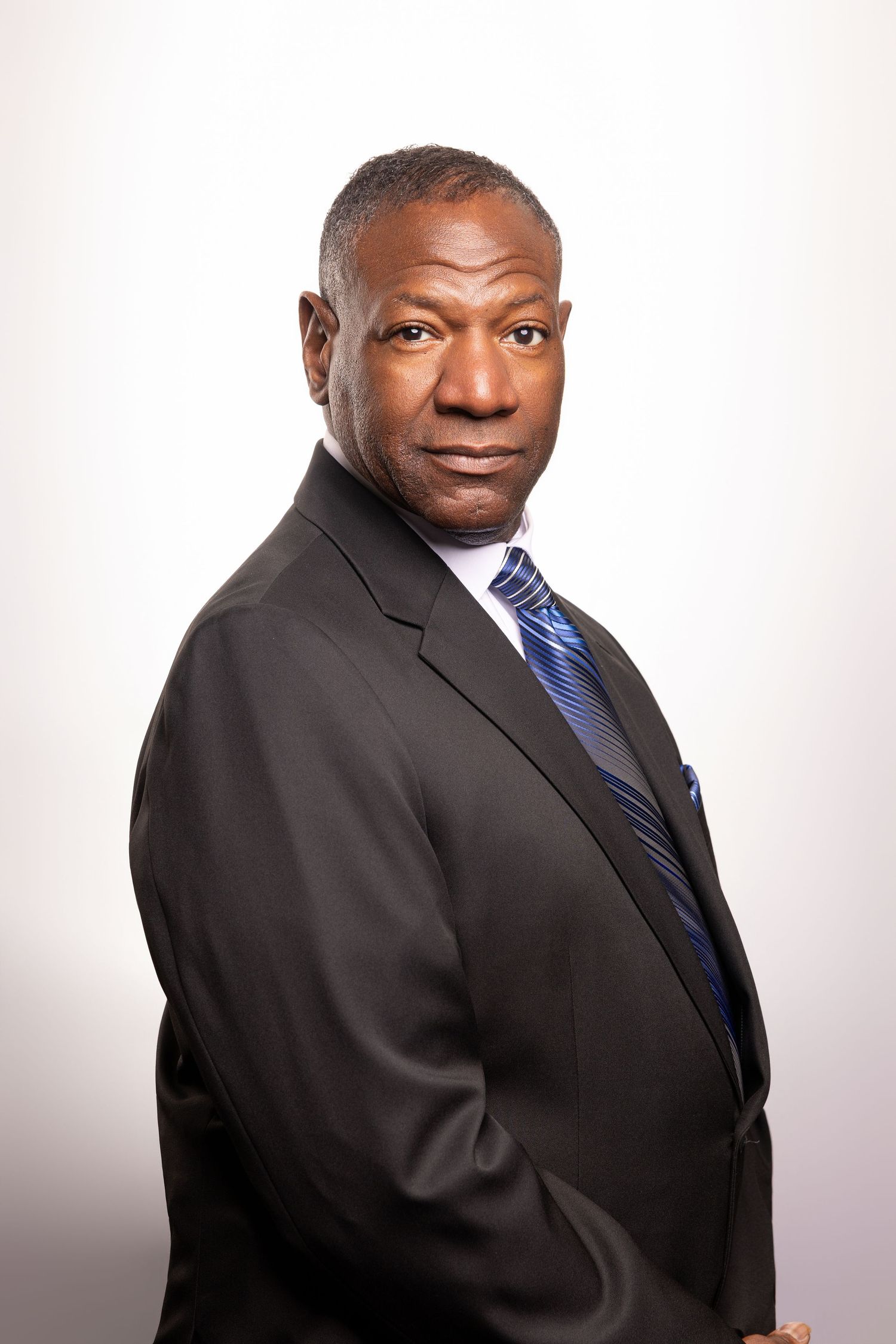 Professional African American businessman in dark suit and blue striped tie posing confidently against white background.