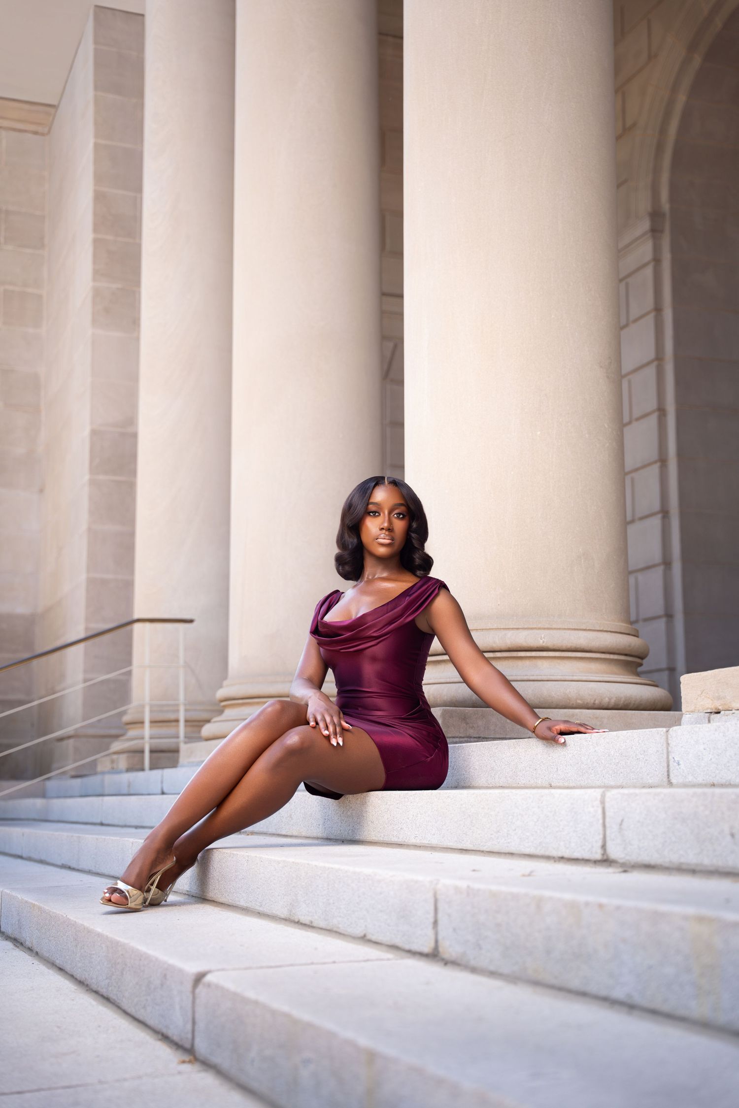 Young woman in burgundy mini dress poses elegantly on marble steps beside classical stone columns.