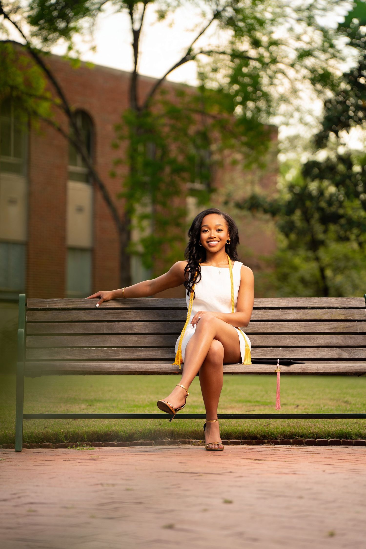 Young woman in white dress sitting on a bench outdoors, smiling, holding graduation cap and tassel.