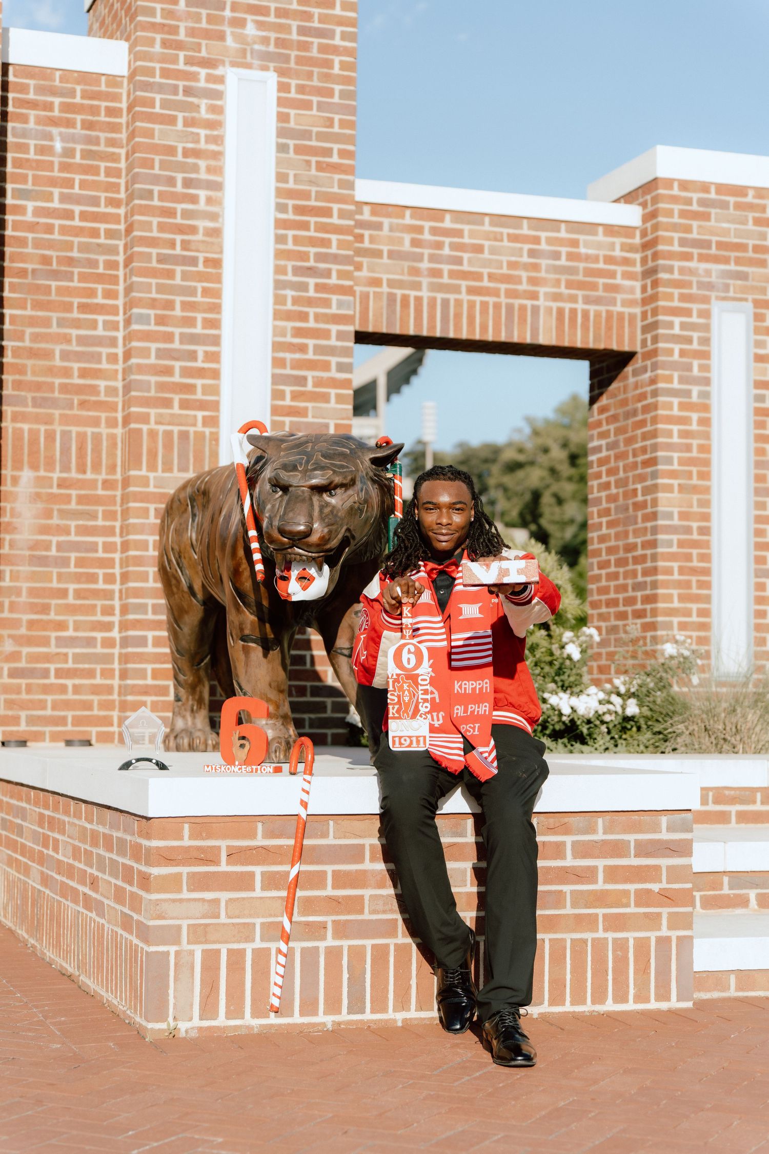 Young man in red and black attire poses beside a bronze tiger statue at a brick university entrance.