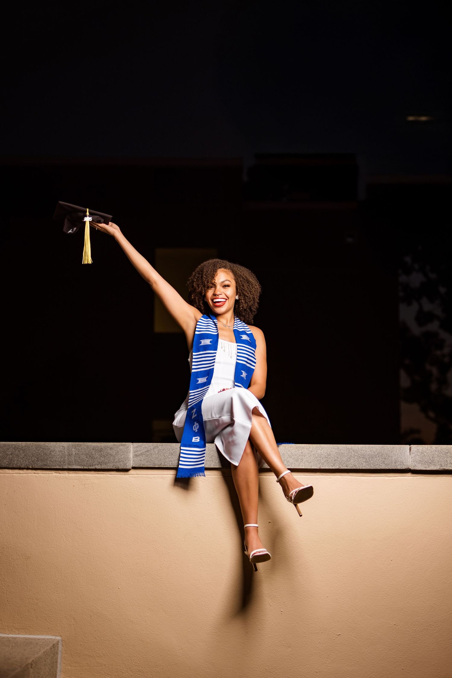 Graduate in blue stole holds up tassel while sitting on ledge.