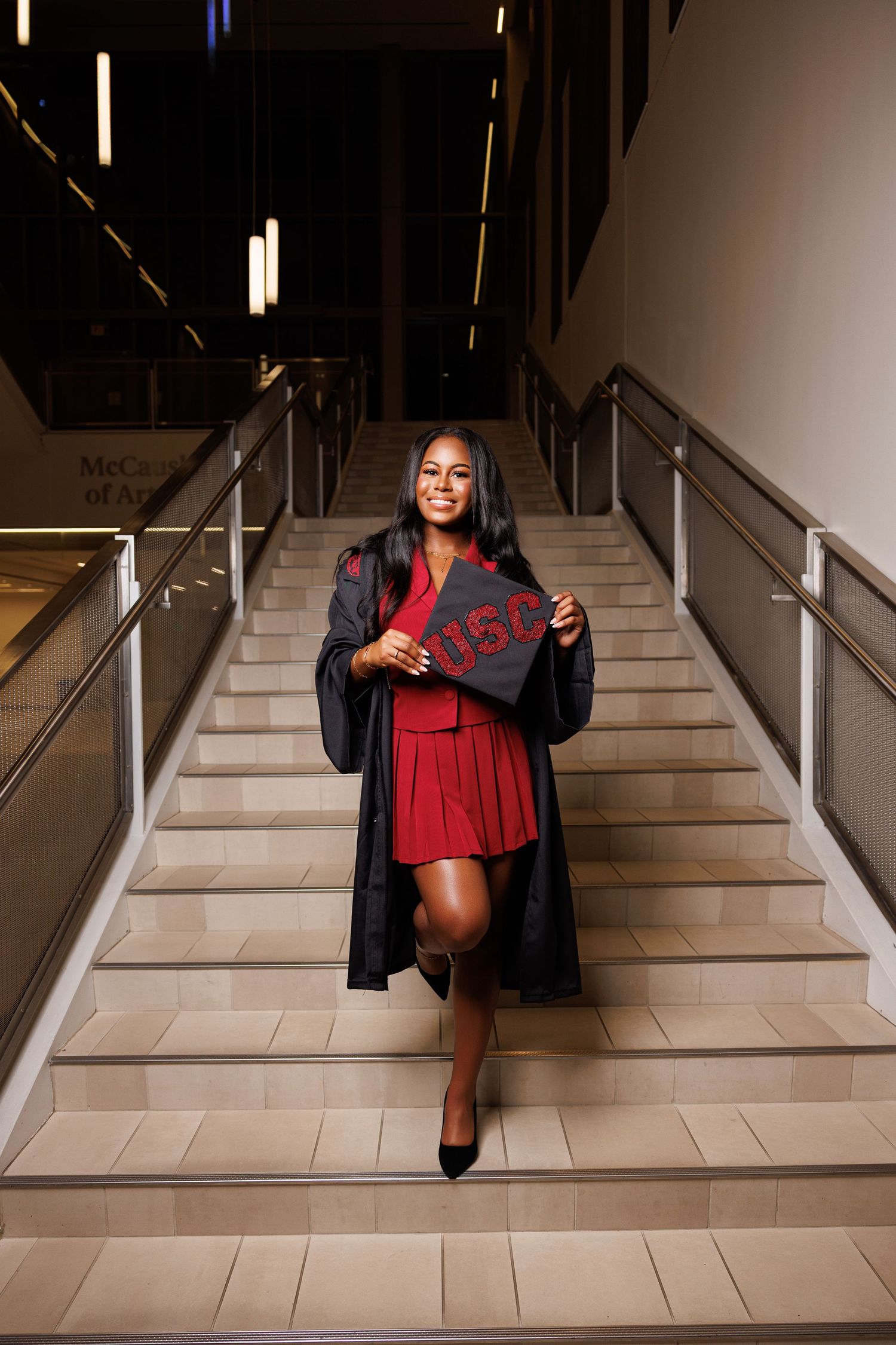 Graduate in red dress and black gown poses on grand staircase.