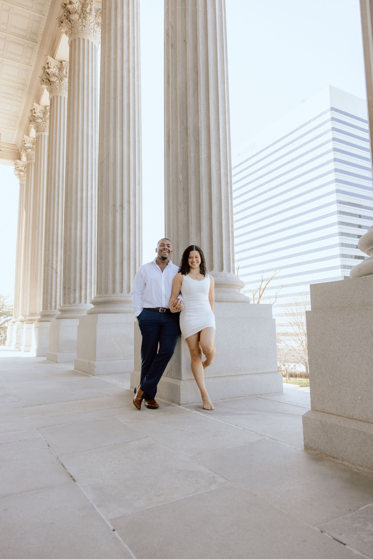 Elegantly dressed couple poses between classical columns of a grand building.