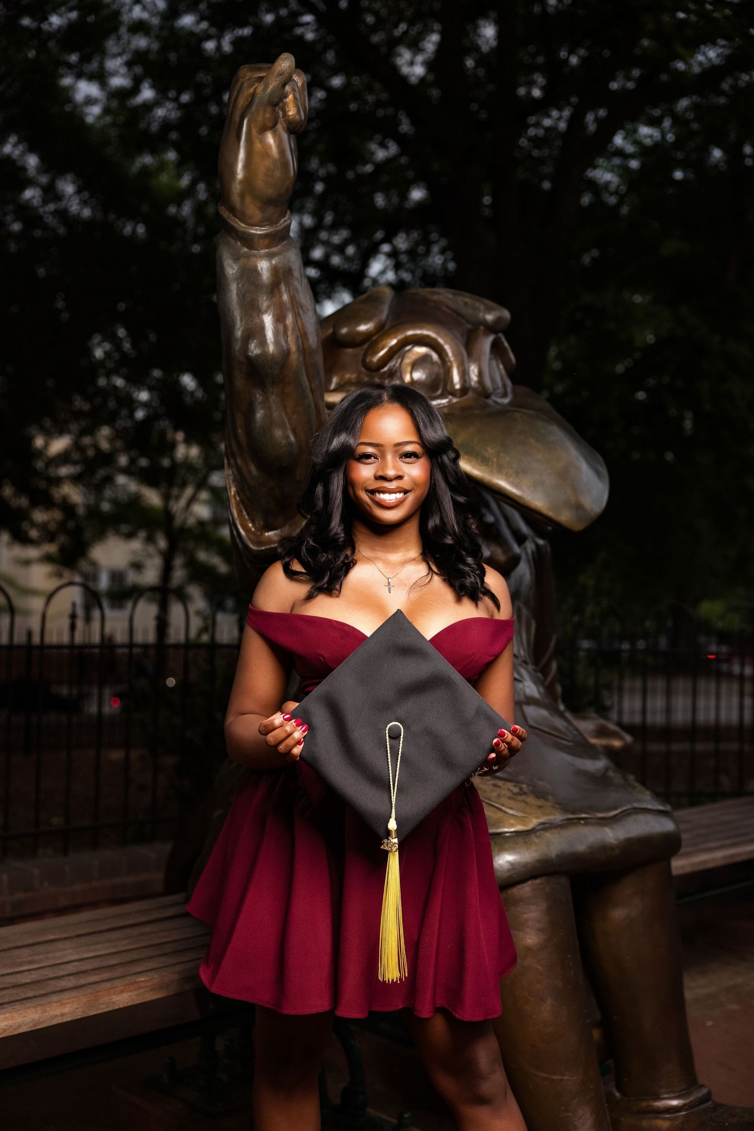 Graduate poses joyfully with a playful university statue in a burgundy dress during commencement photos.