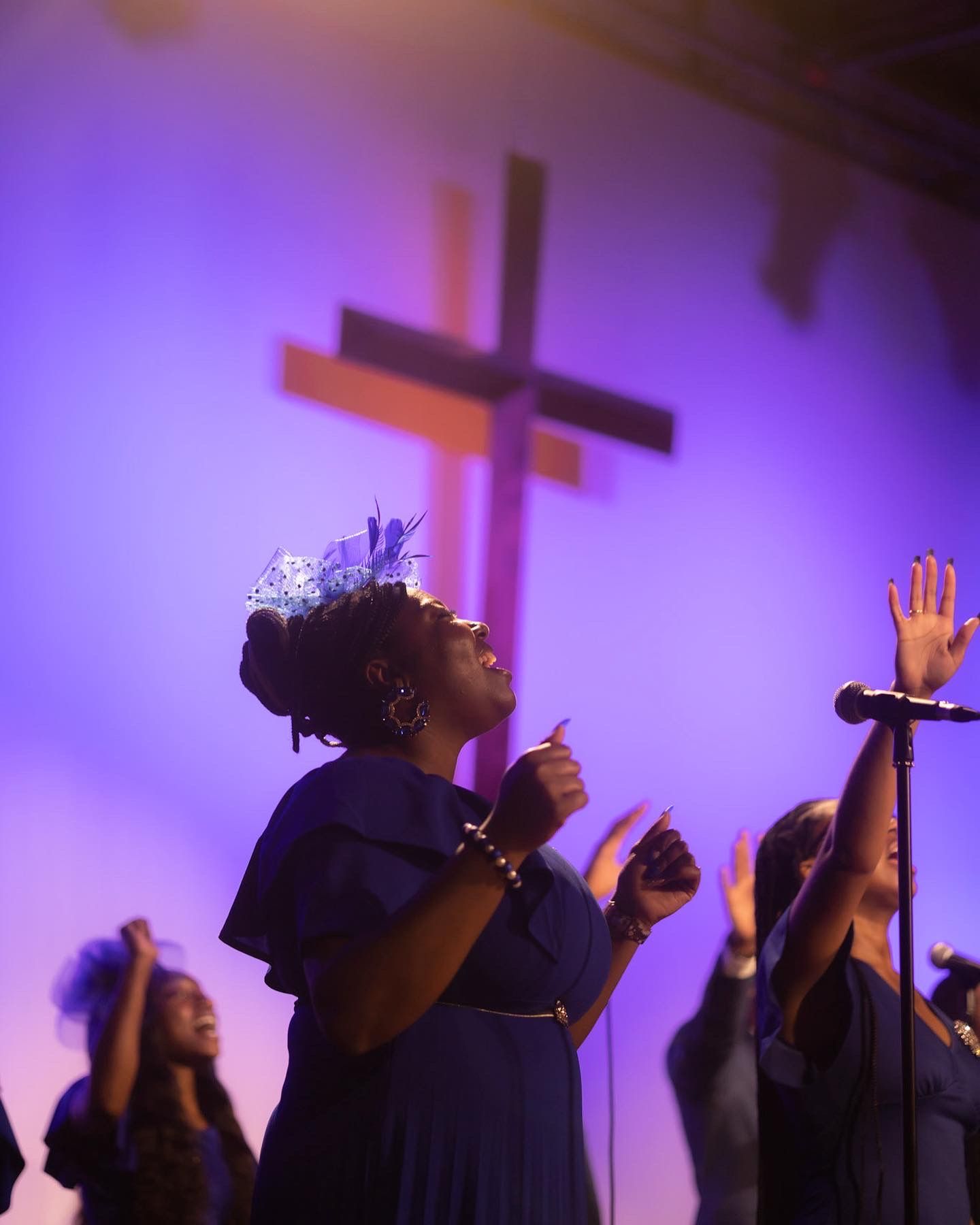 Silhouettes of choir members singing with raised hands against purple-lit backdrop with cross.