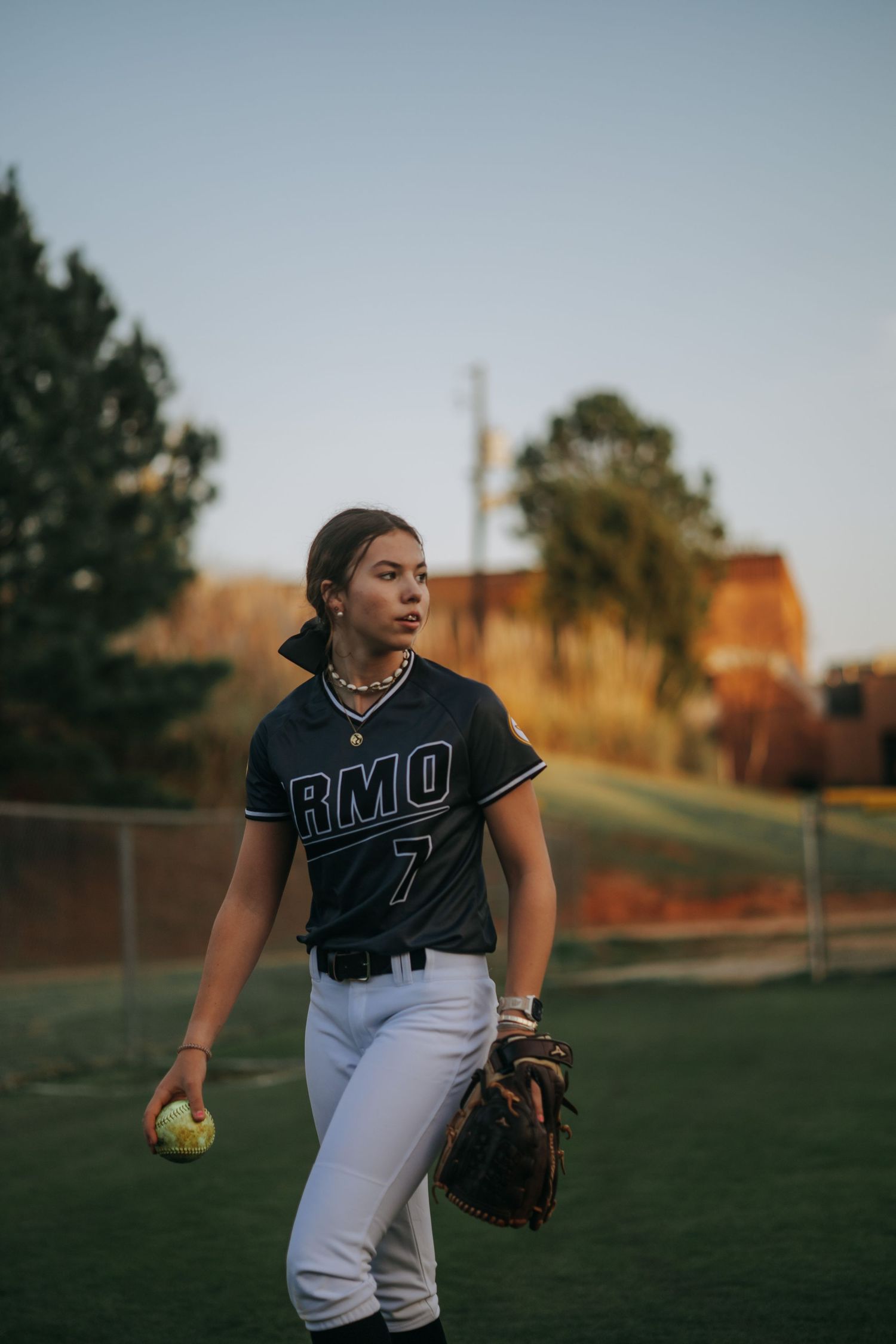 Softball player in RMO jersey holds ball and glove on field at sunset.