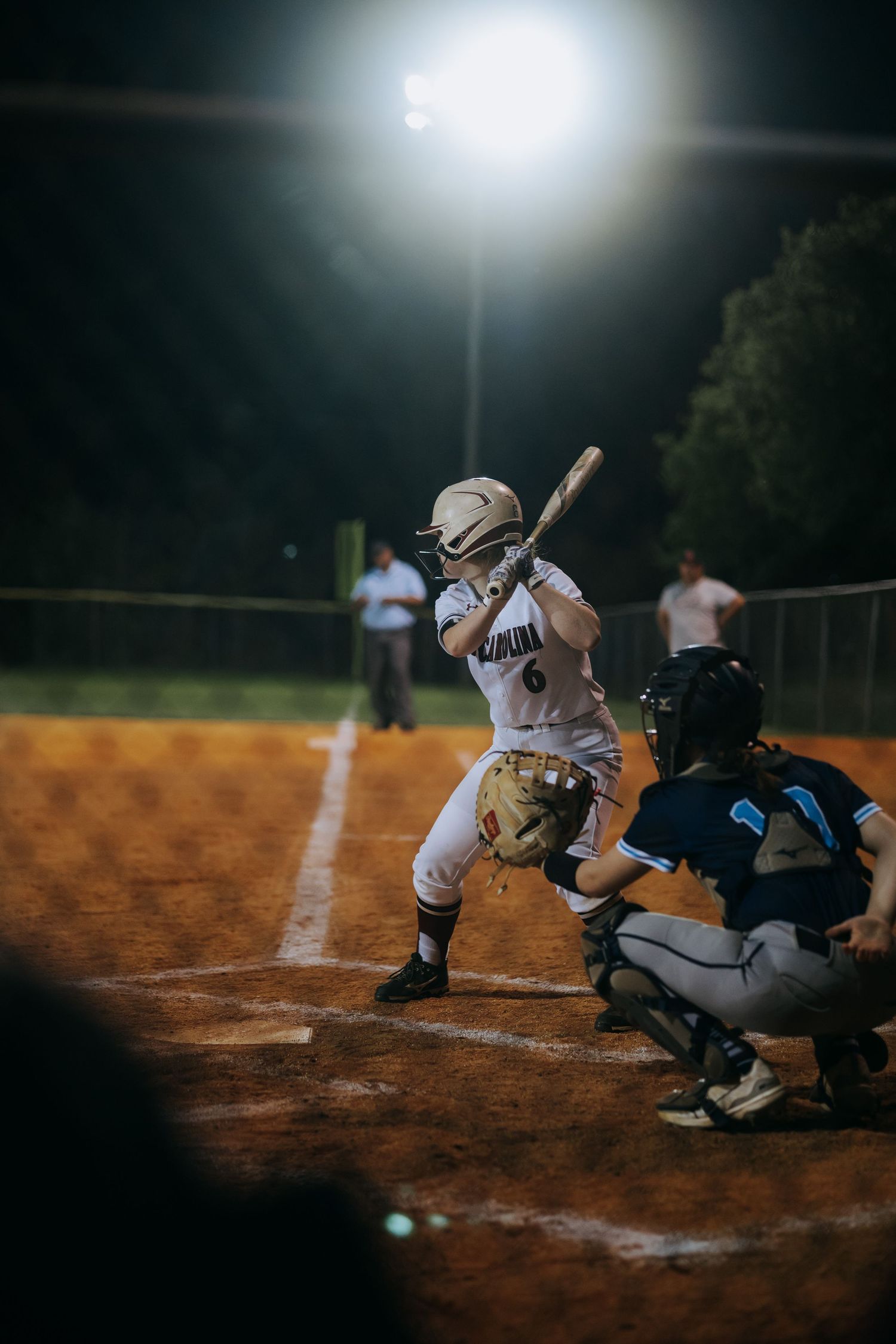Softball player at bat during nighttime game with catcher and umpire visible on dirt field under stadium lights.