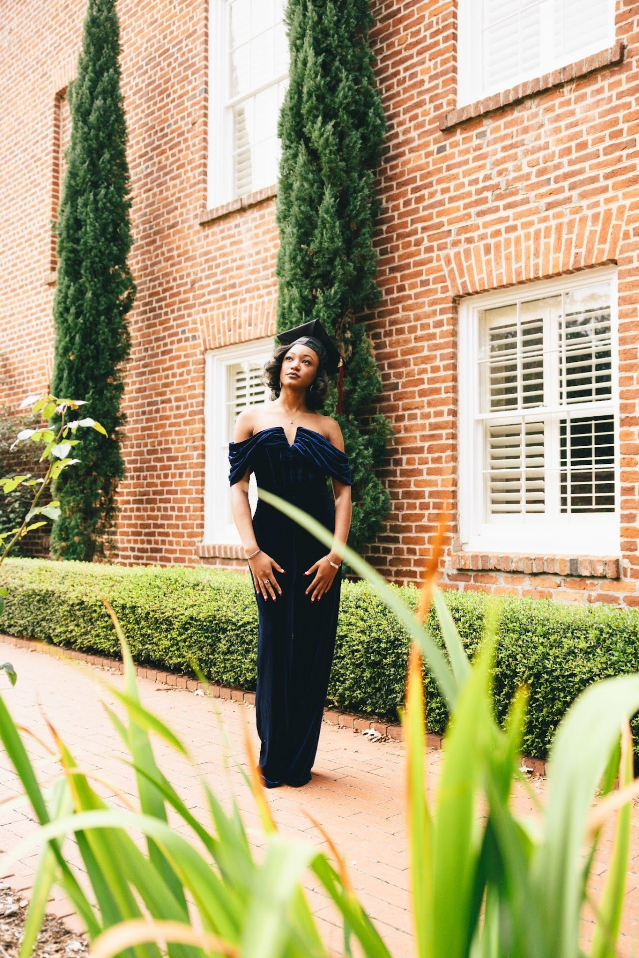 Graduate in black cap and gown takes photos in front of a brick building with landscaped gardens and windows.