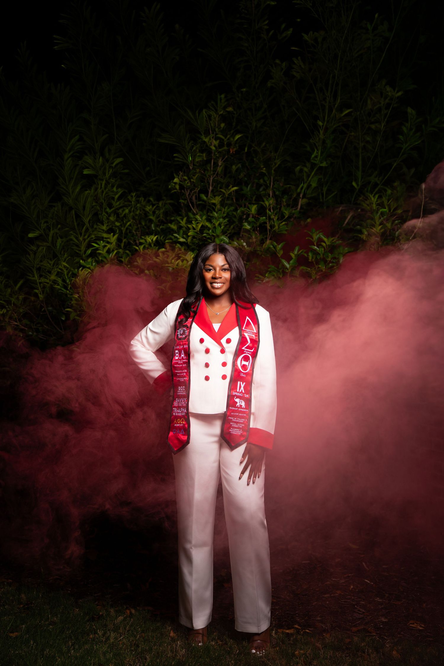 Young woman in white suit with red graduation stole poses amid red smoke at night.