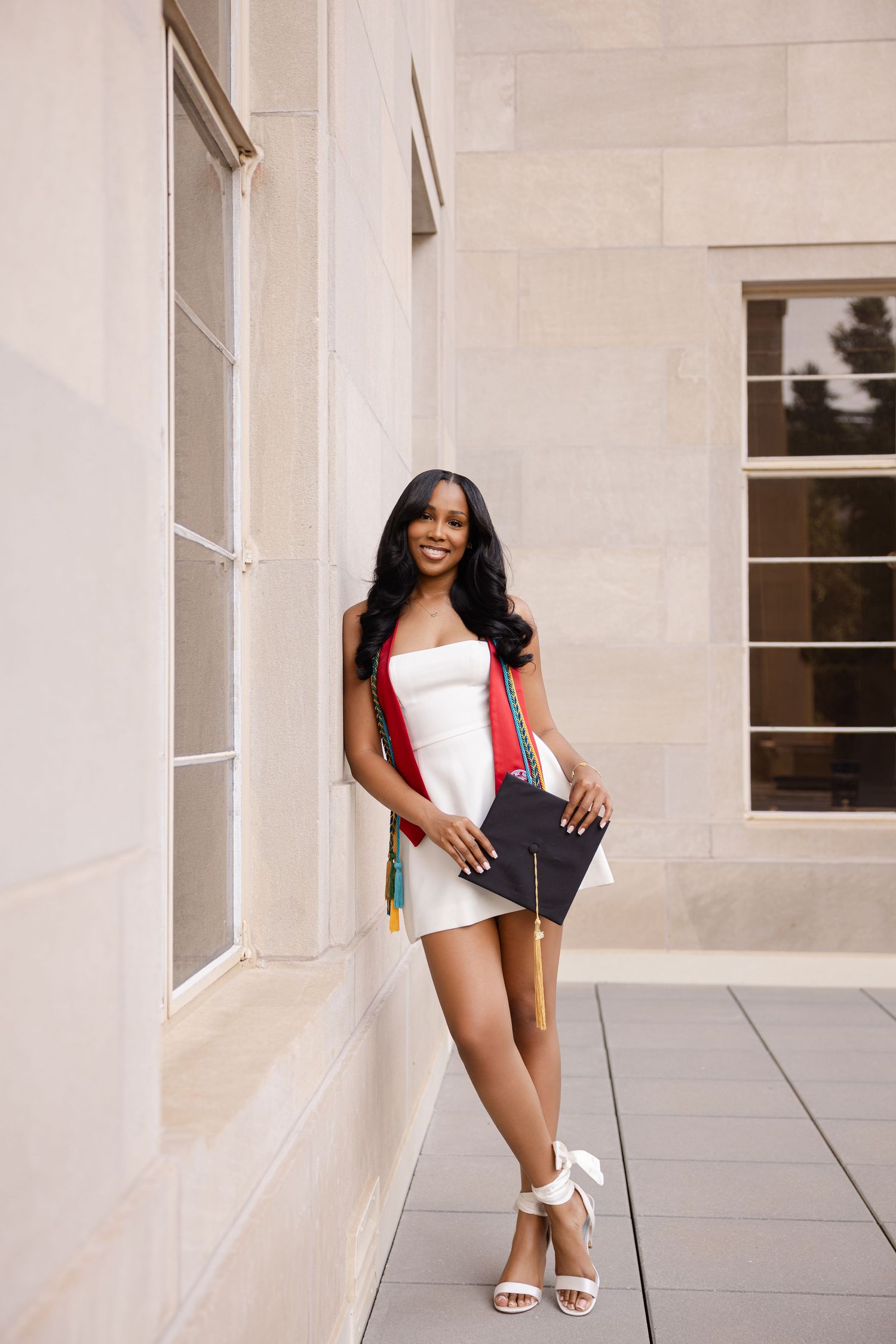 Young woman in white dress holding graduation cap and colorful tassel, posing confidently outdoors near a building.