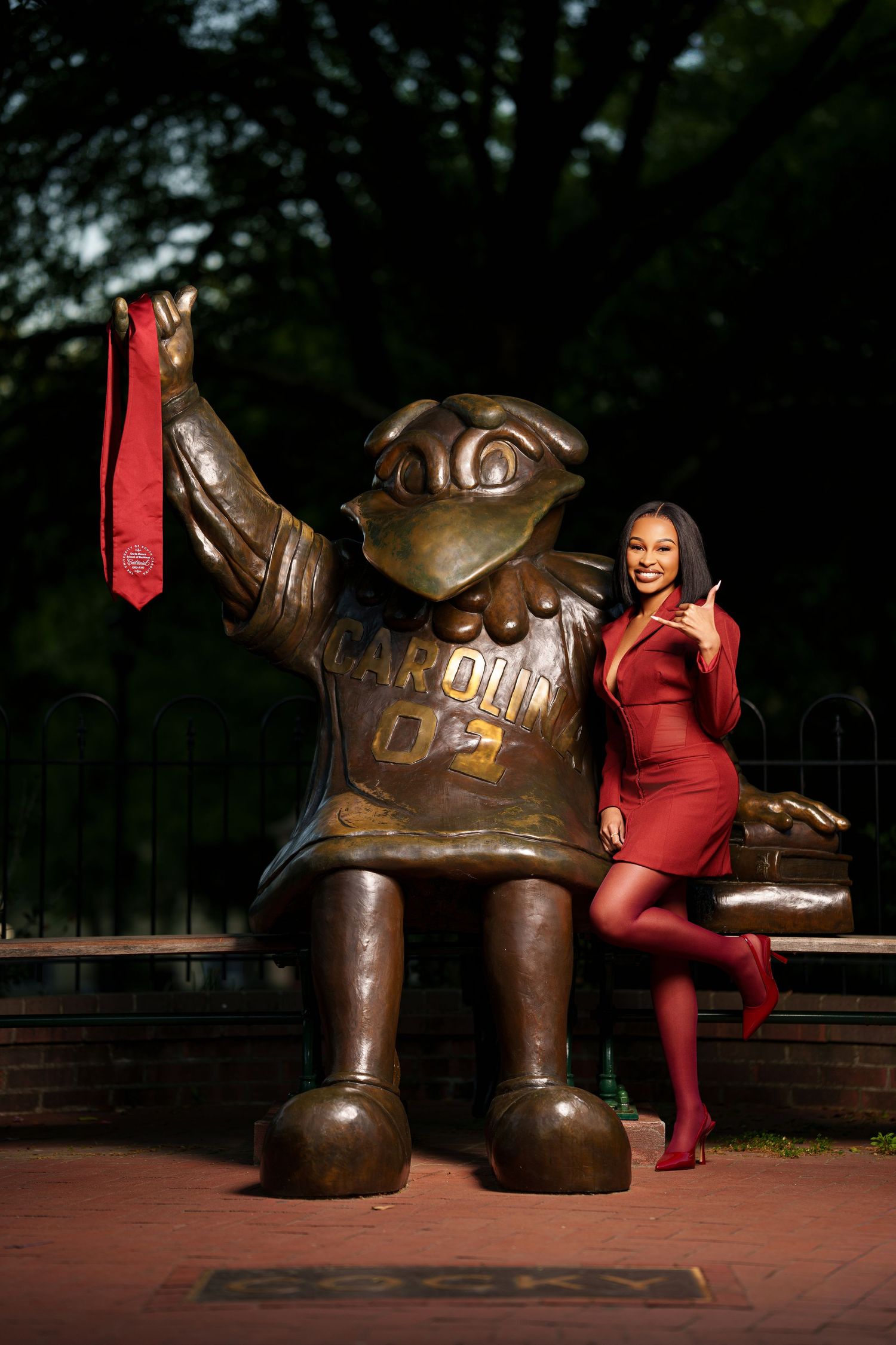 Young woman in red dress poses beside University of South Carolina Cocky mascot bronze statue on campus.