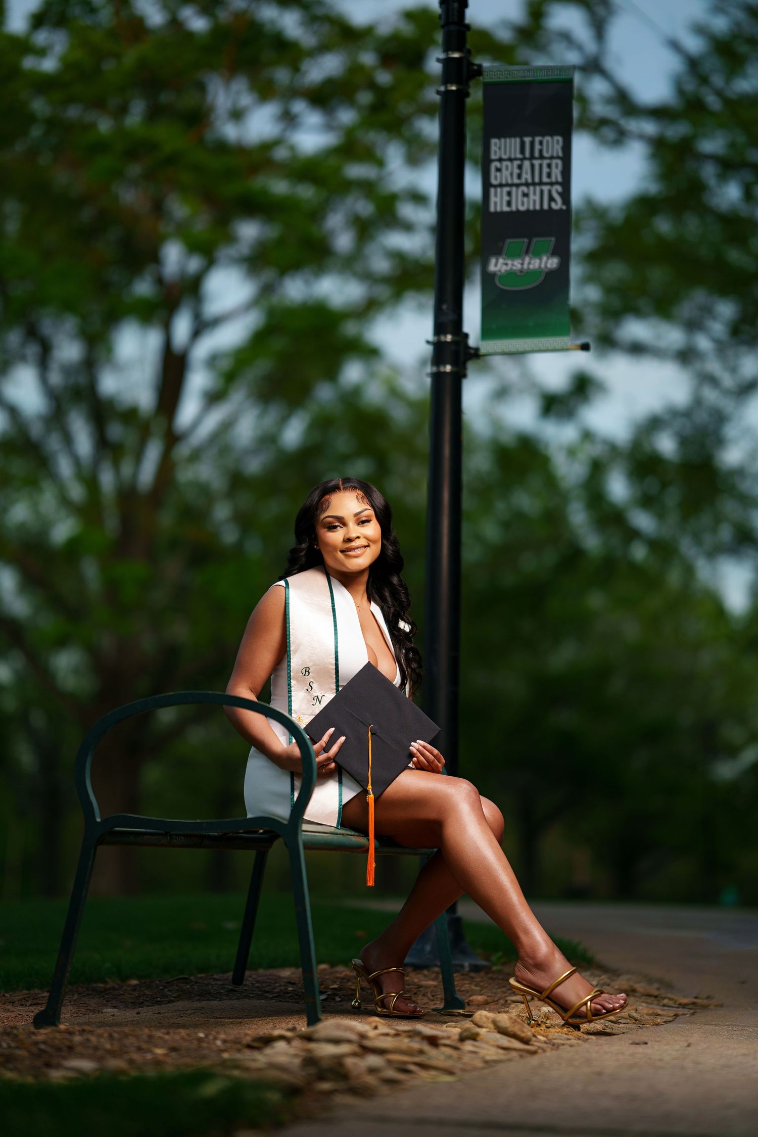 Young woman in graduation attire sits on a bench holding her cap near a university lamppost banner outdoors.
