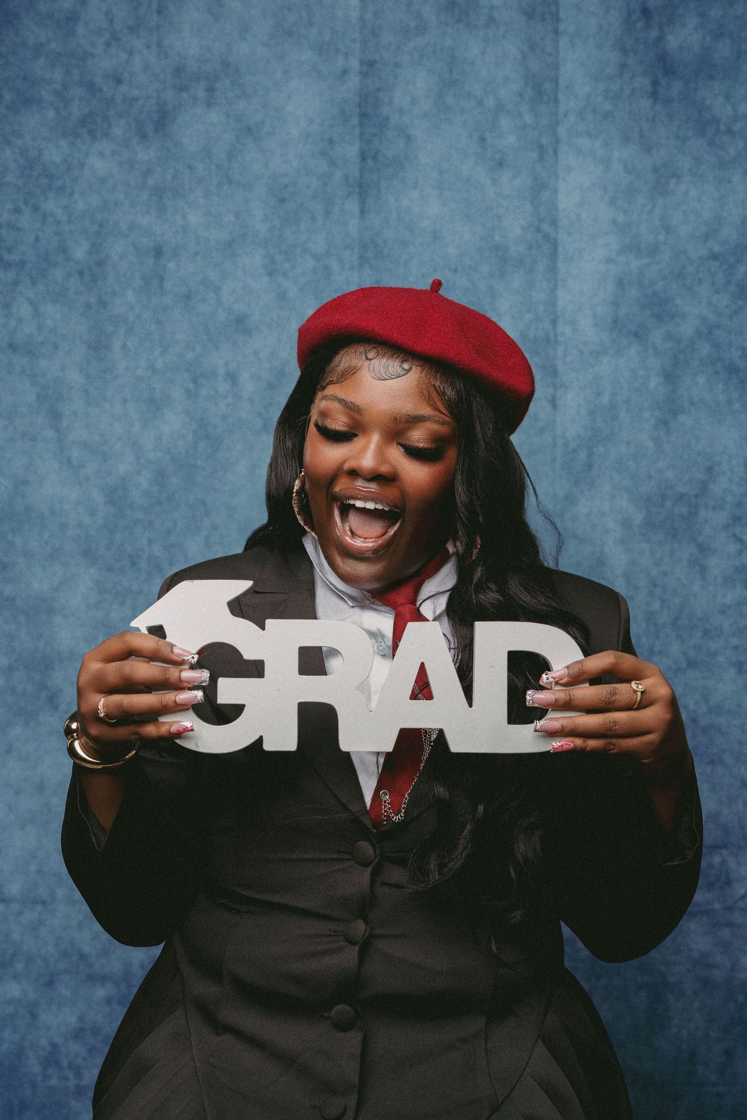 A joyful young woman wearing a red beret and dark blazer holds a GRAD sign, celebrating her graduation.