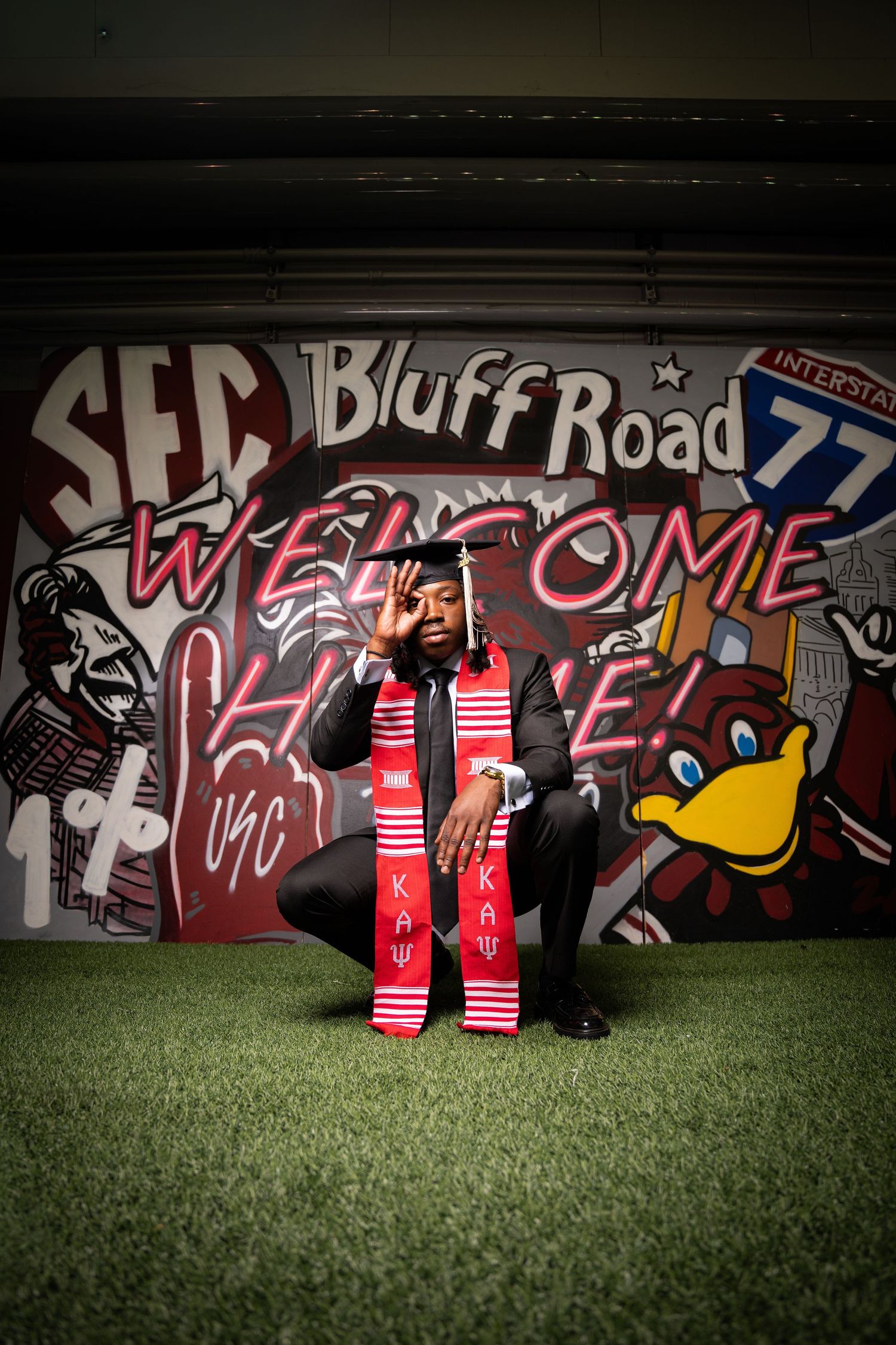 Graduate in cap and gown with Kappa stole poses in front of colorful USC Bluff Road mural at graduation.