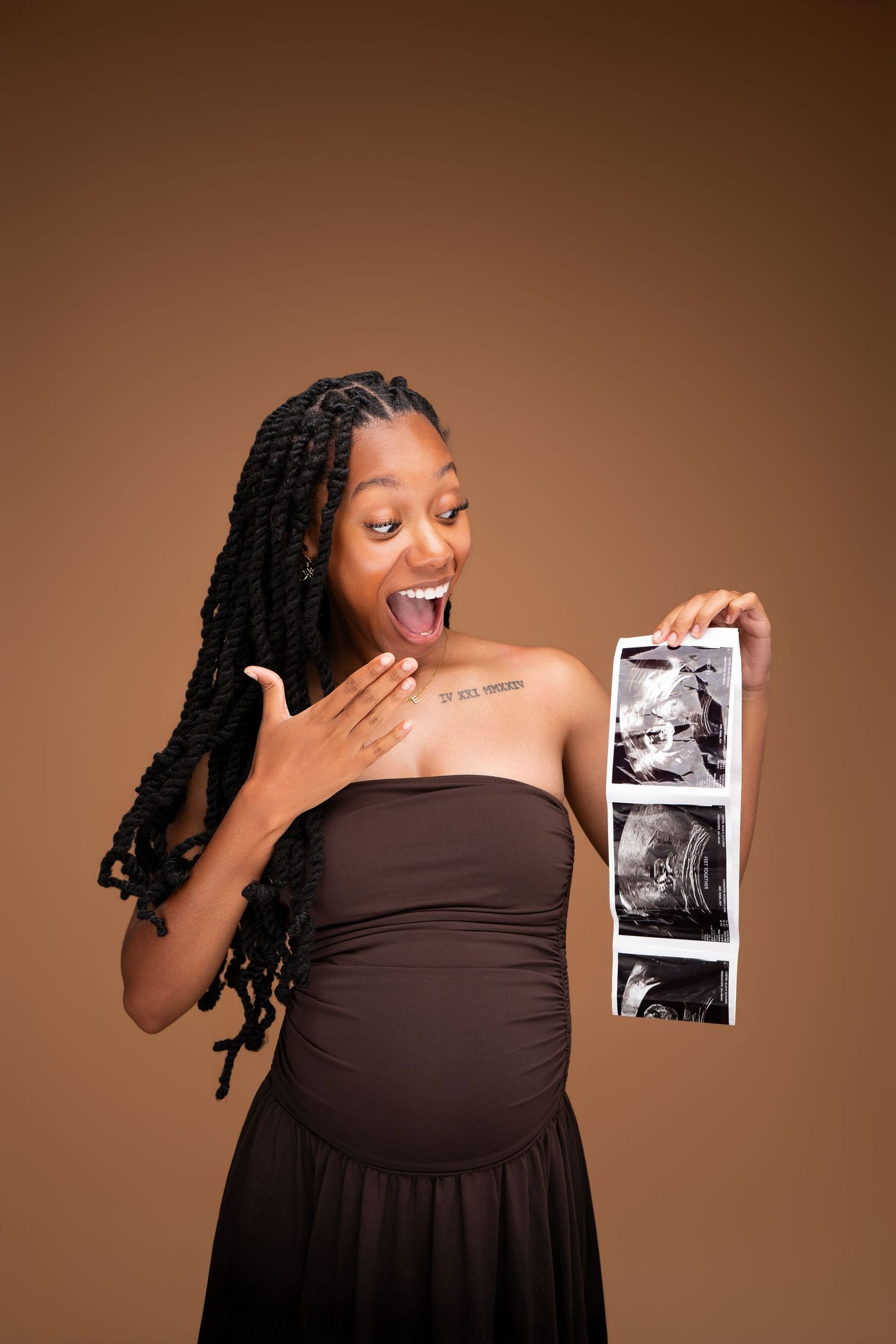 Excited pregnant Black woman in brown dress holding ultrasound images, showing joyful reaction on brown background.