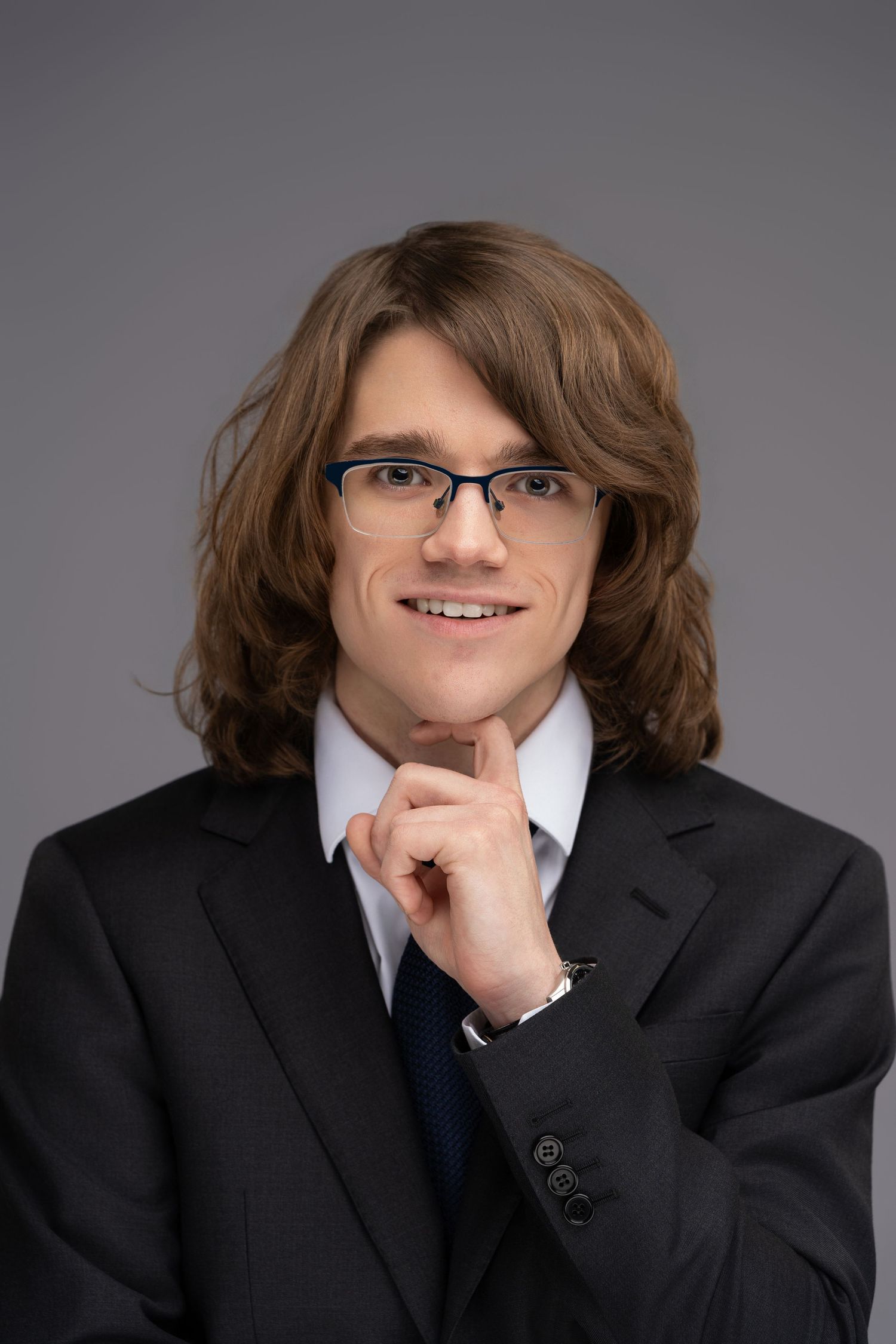 Young professional man with long brown hair and glasses in a black suit, smiling confidently against gray background.